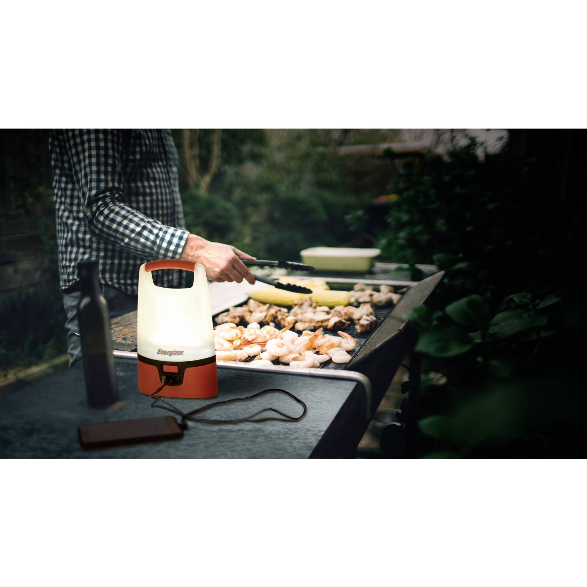 A person is grilling chicken and vegetables outdoors at dusk. A lantern illuminates the barbecue area.