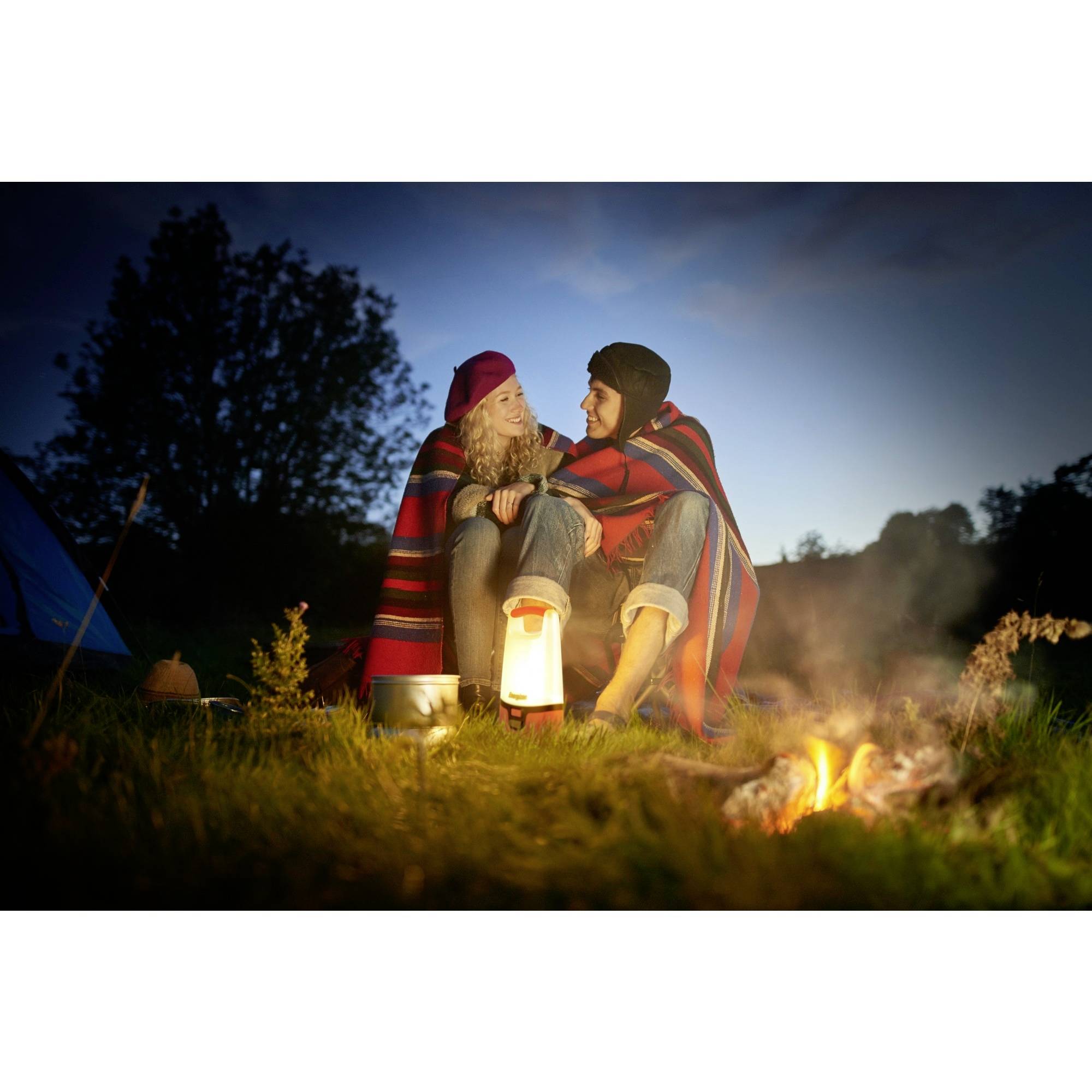 Two people sit wrapped in blankets beside a campfire on a meadow. It is twilight, with a tent in the background.