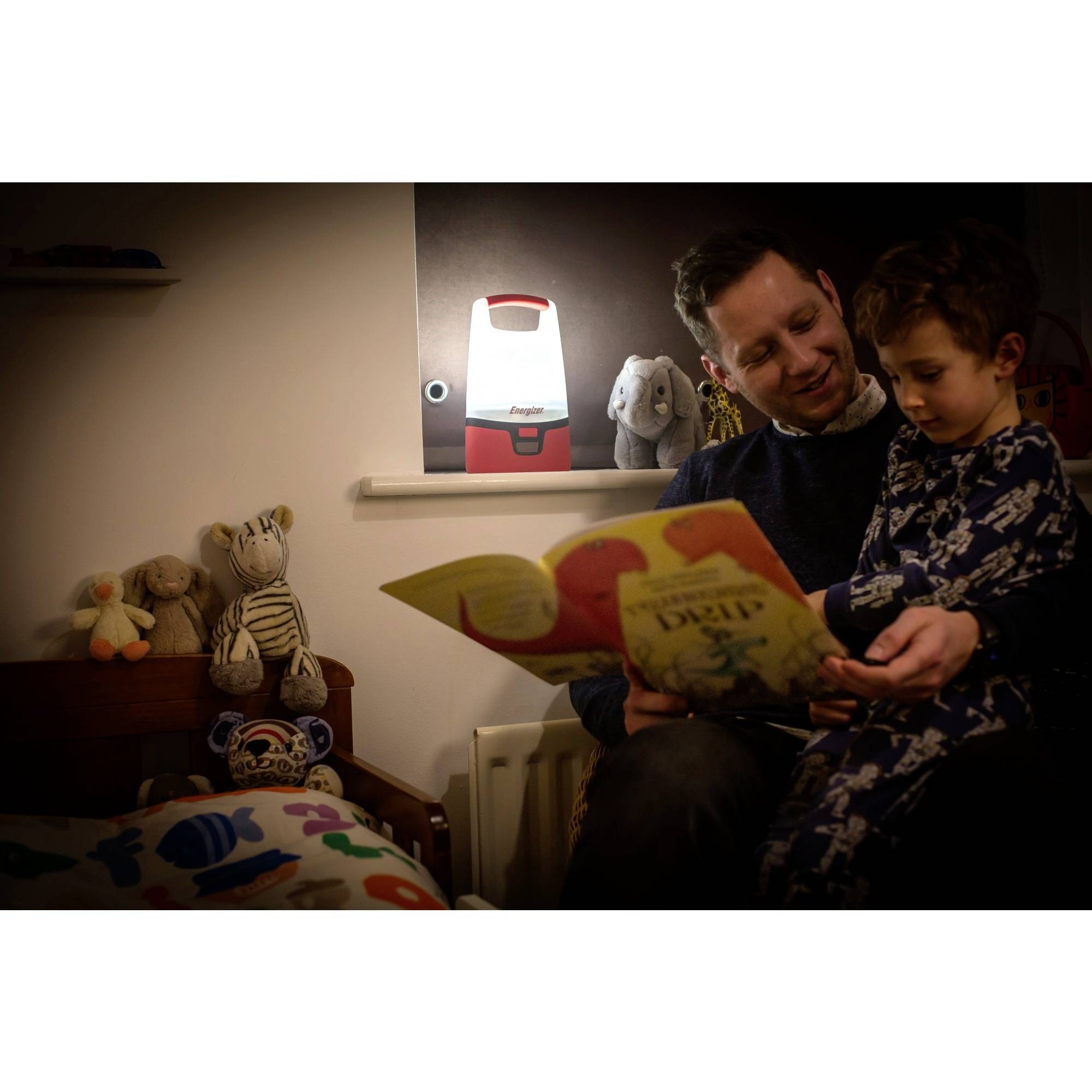 A father is reading a story to his child in a cosy, dimly lit bedroom. Soft toys are in the background.
