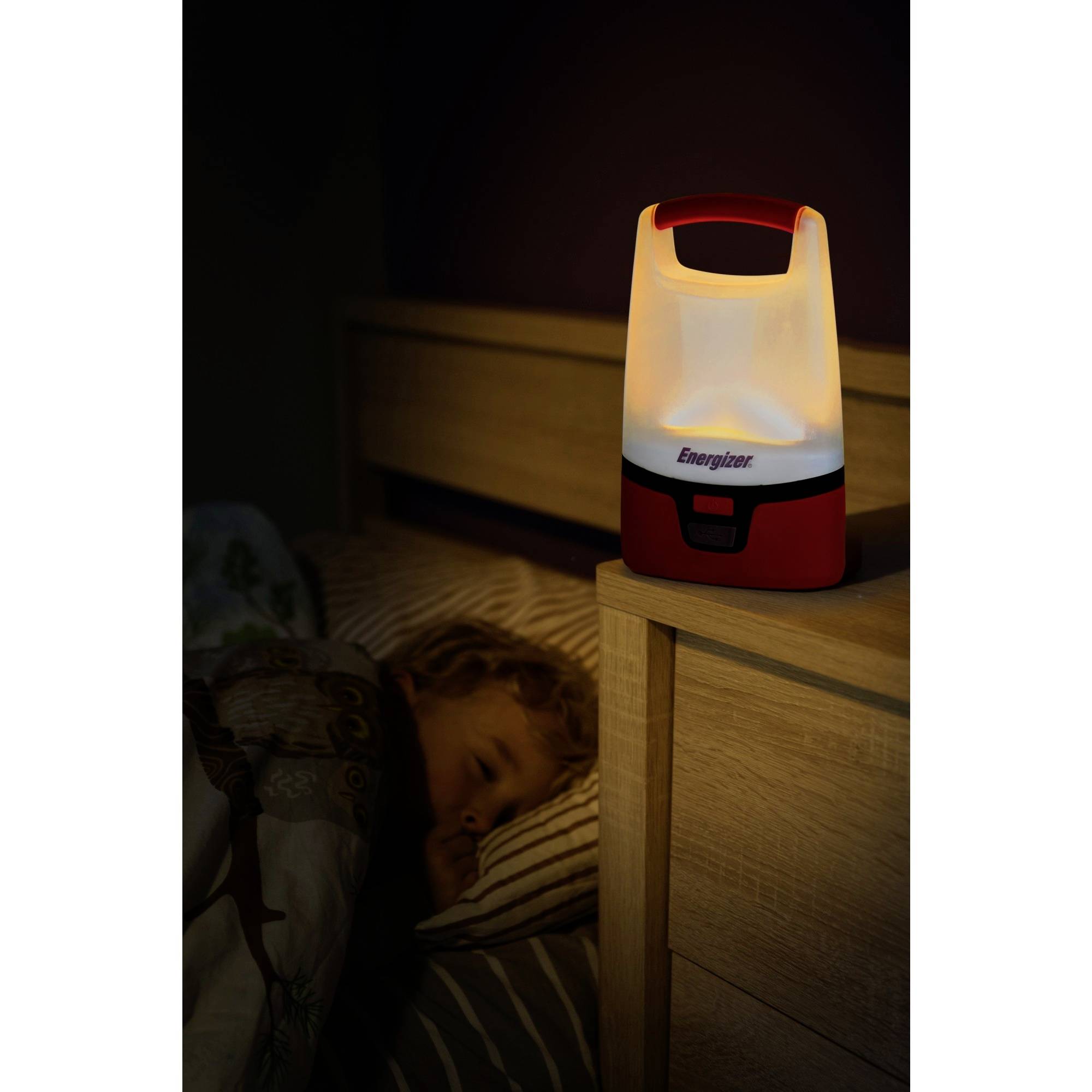 A glowing camping lamp sits on a bedside table next to a sleeping child in a dark bedroom.