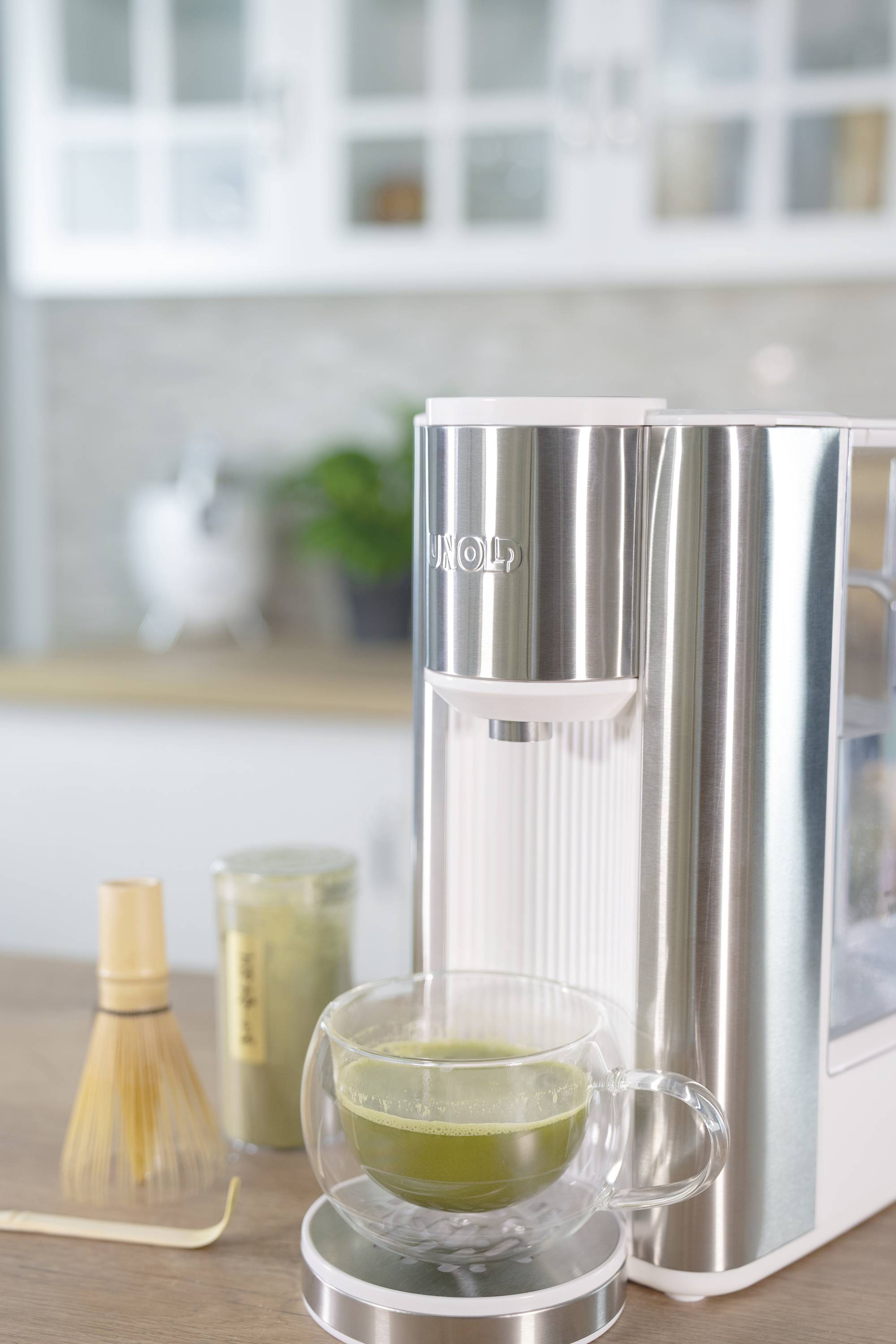 A stainless steel machine for preparing matcha stands on a kitchen worktop. Beside it is a cup of matcha and a traditional bamboo whisk.