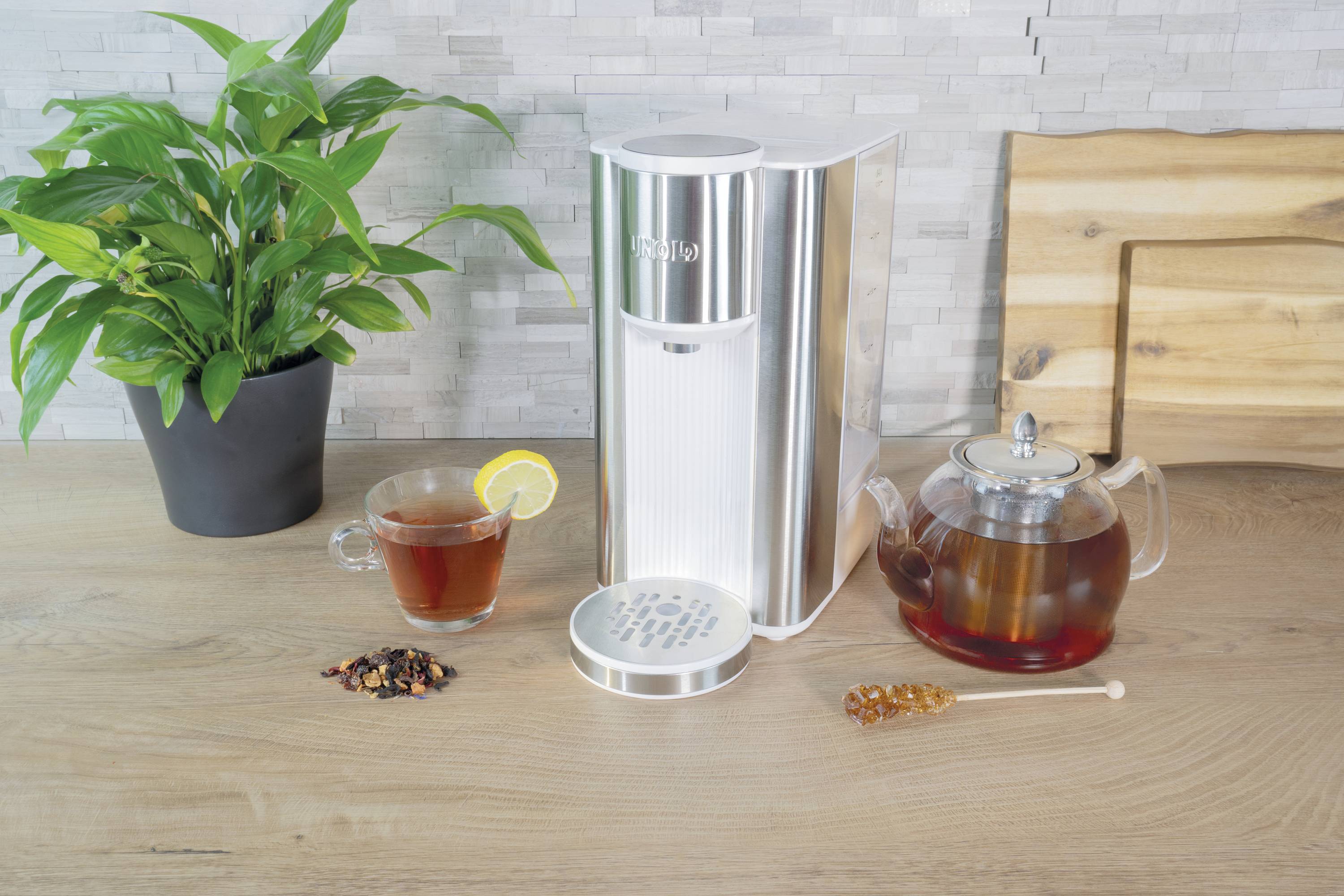 A silver sparkling water maker on a kitchen worktop next to a cup of tea with lemon, a kettle and a plant.
