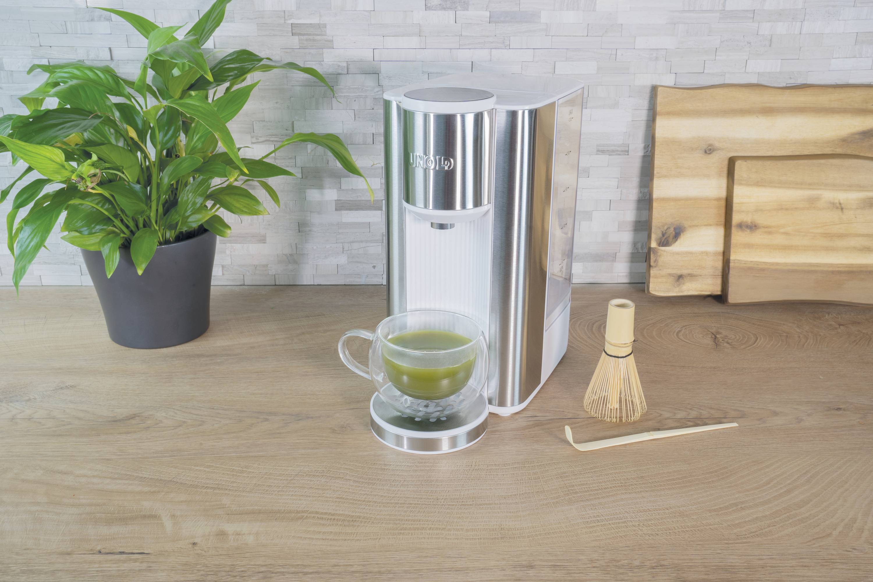 Silver teapot with a cup of green tea, alongside a bamboo whisk and wooden spoon on a wooden table. A potted plant sits to the left.