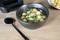 A black bowl with clear soup, floating bread croutons, spring onions and herbs. Beside it lies a black spoon.