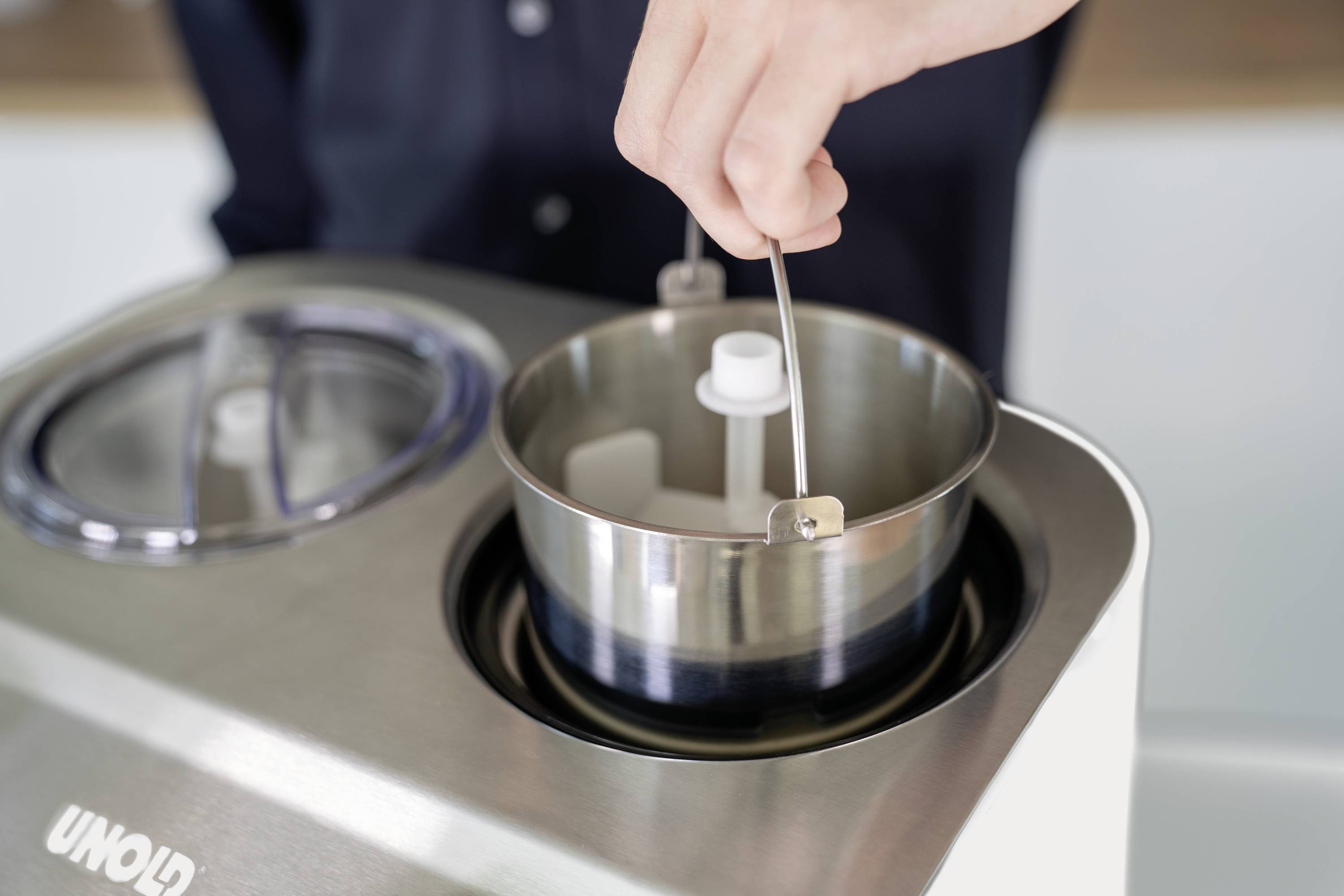 A hand holds the mixing paddle of an ice cream maker over the open stainless steel bowl of the machine.