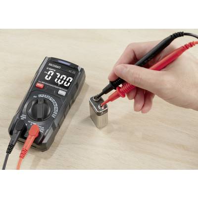 'Hand with test probes of a multimeter measuring the voltage of a 9-volt battery. Multimeter shows 7.00 volts. Wooden surface in the background.'