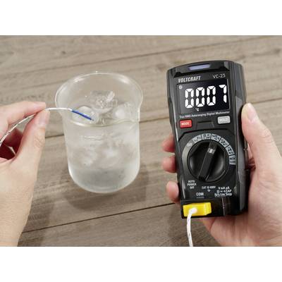 A person is measuring the water temperature in a beaker using a digital multimeter, which displays 0.07 °C.
