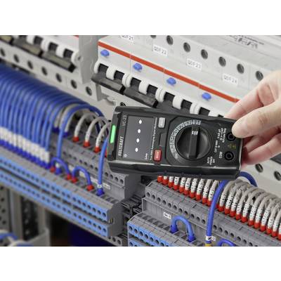 A hand holds a multimeter in front of an open electrical cabinet with blue and grey cables, measuring electrical values.