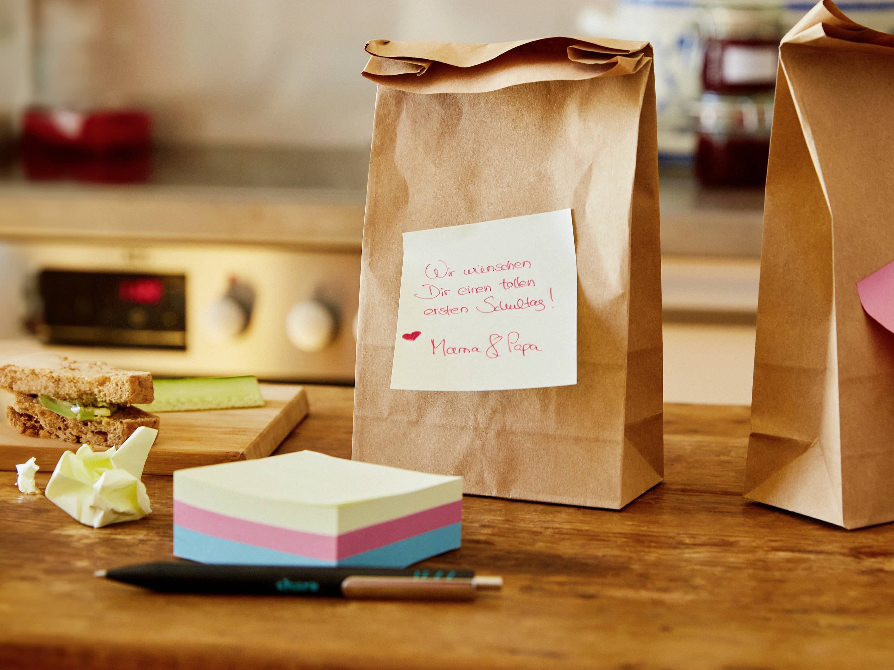 Paper bags with a note reading 'We wish you a fantastic first day at school! Mum & Dad' on a kitchen worktop, with a sandwich and notepad beside them.