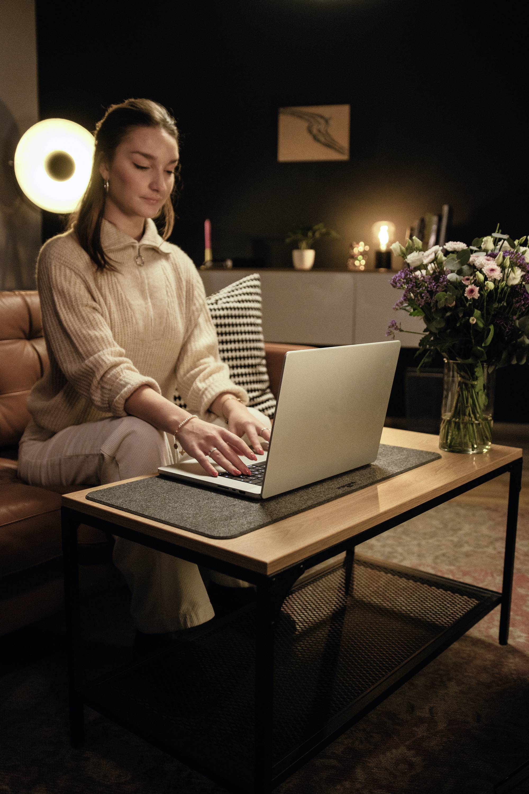 A woman is sitting on a sofa and typing on a laptop; a bouquet of flowers sits on the table beside her. The living room has dimmed lighting in the background.