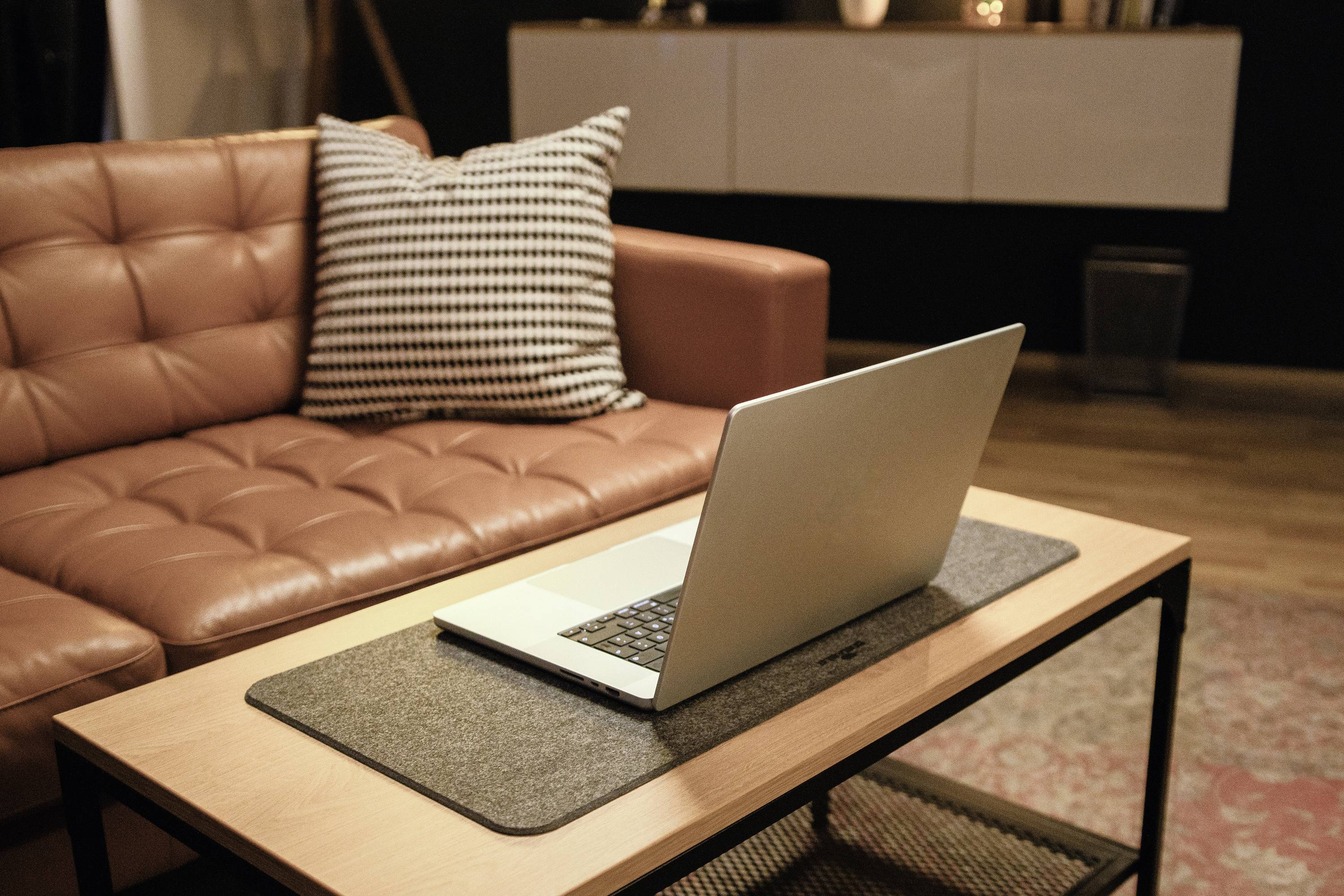 A laptop is sitting on a table in the living room in front of a brown leather sofa with a black and white checked cushion.