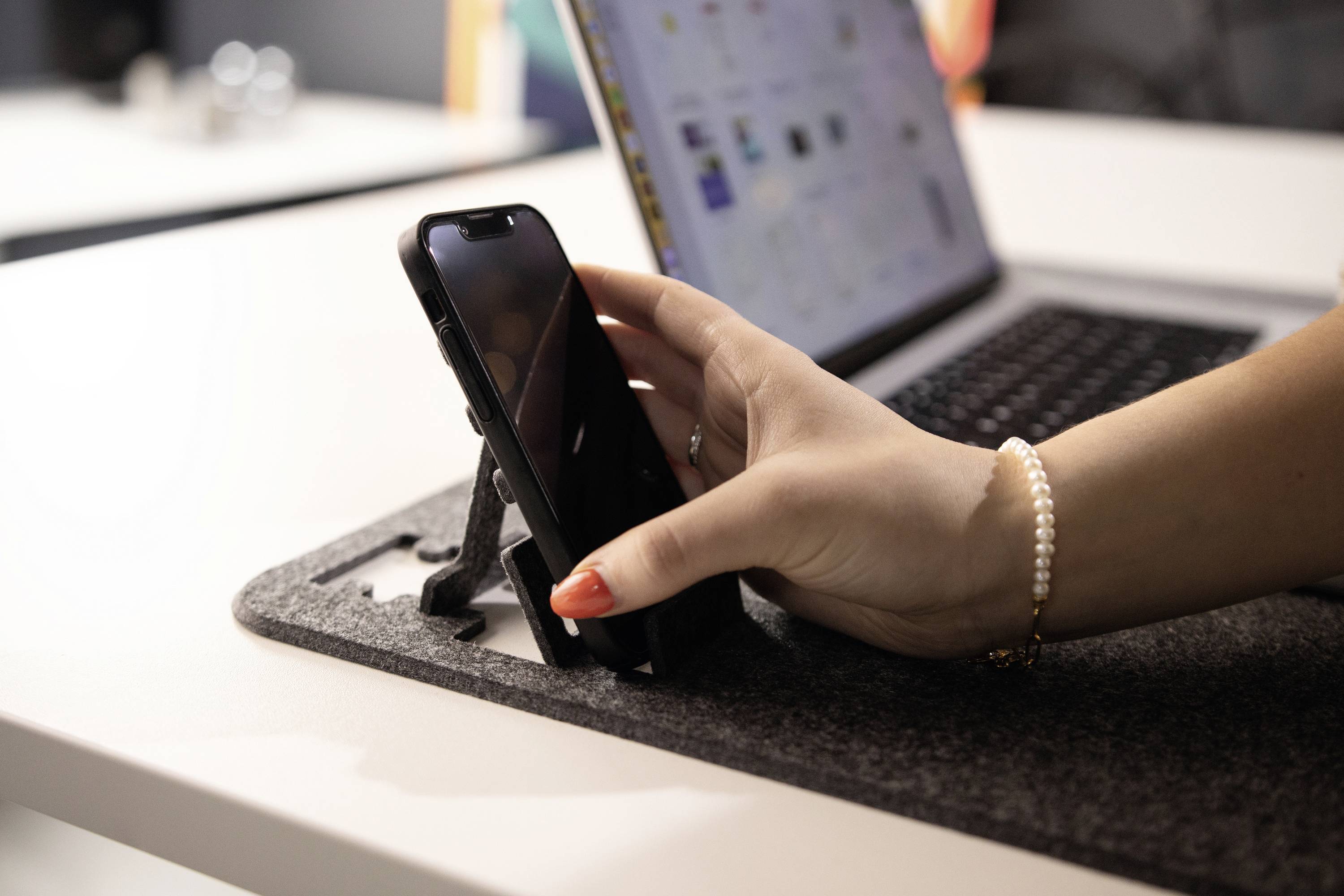 A hand with red nail polish is holding a mobile phone on a table next to an open laptop.