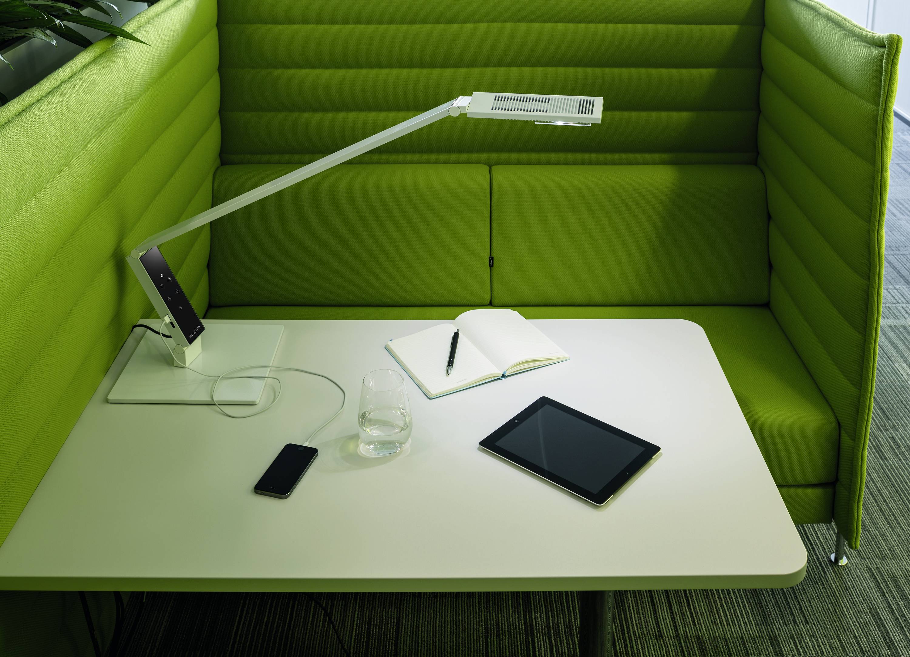 Round table with a tablet, a notebook and a mobile phone. A green upholstered seating area surrounds the table on three sides.