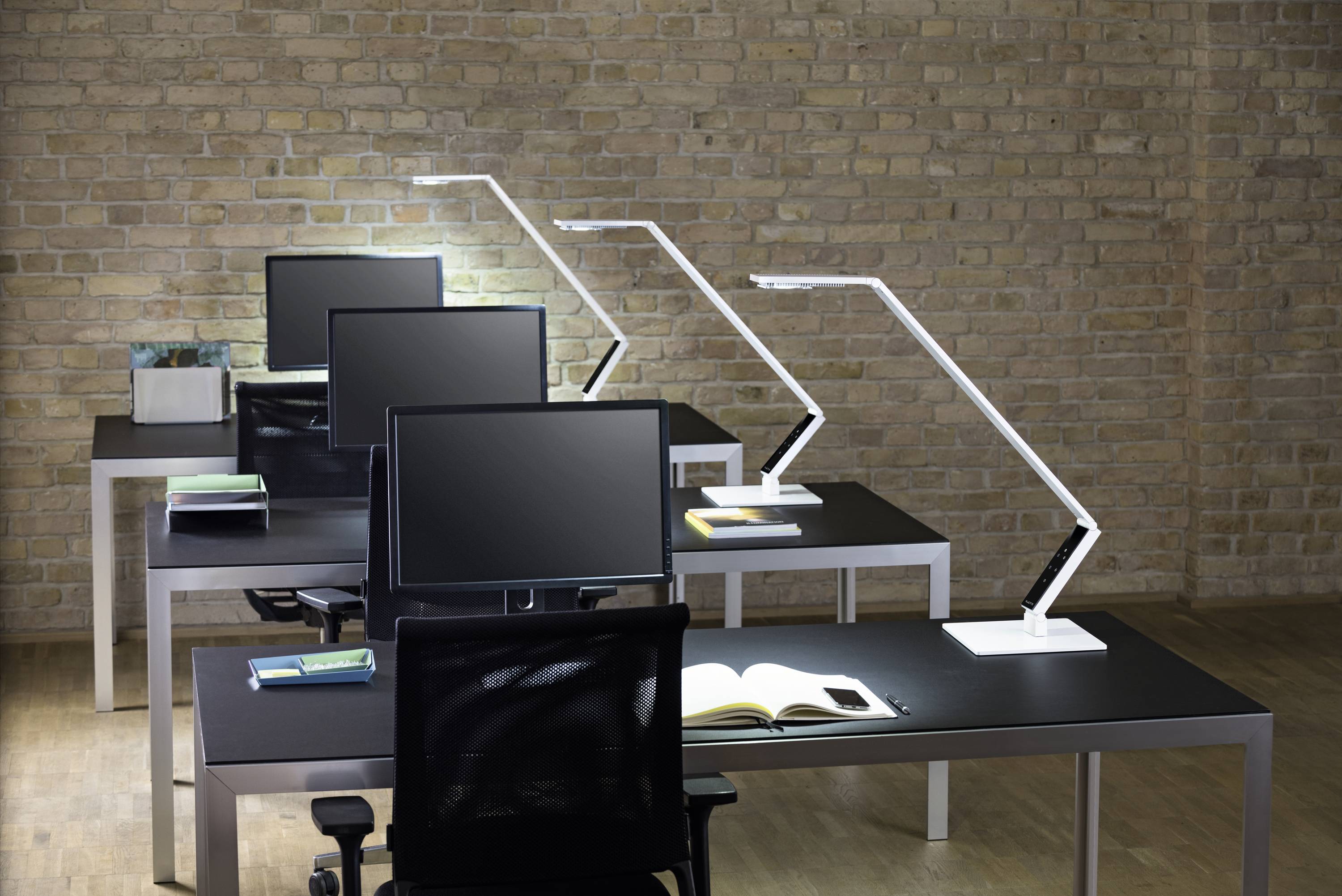 A modern office with four desks arranged in a row. Each desk has a monitor, a desk lamp, and an open book.