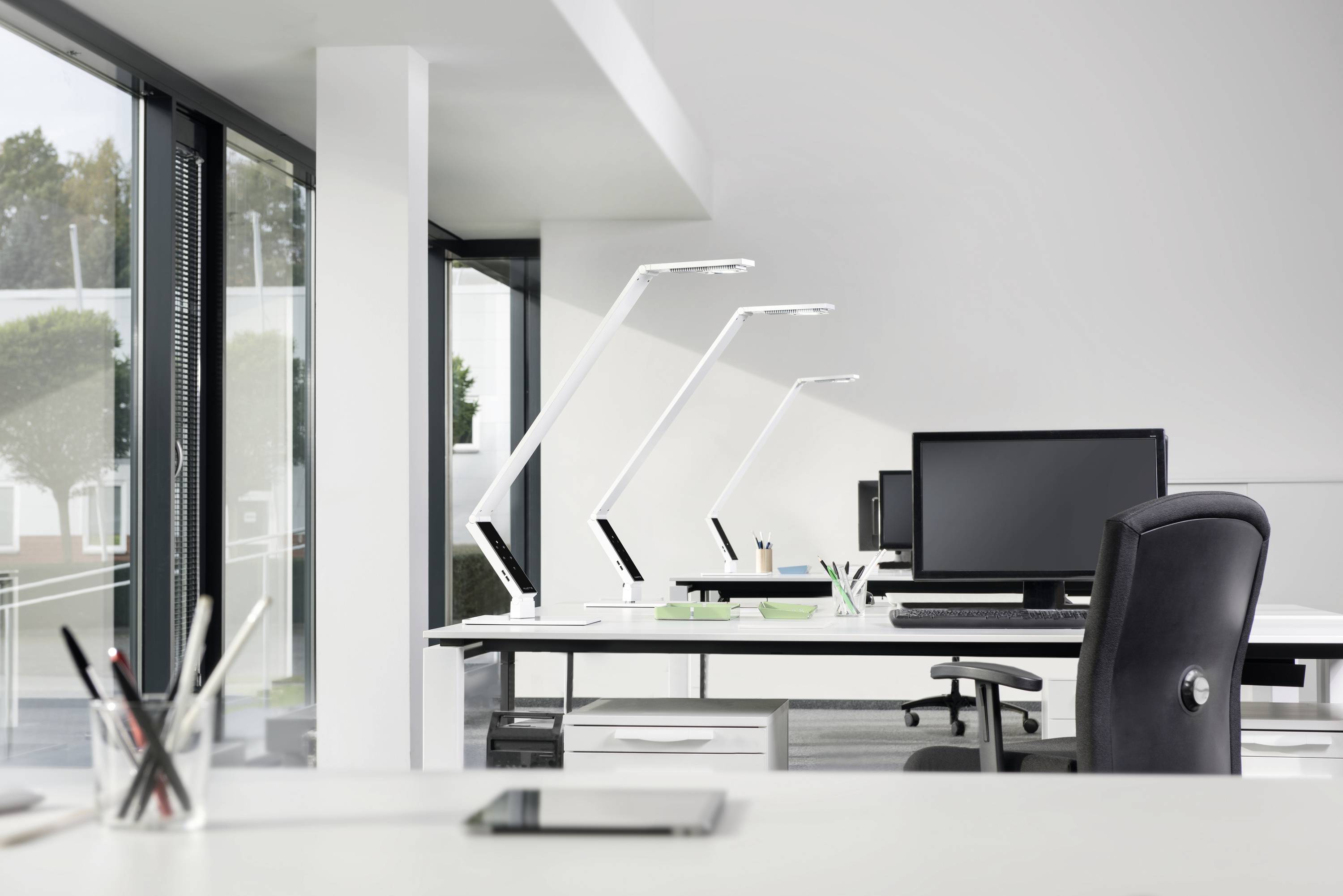 A modern, bright office with desks, black chairs, computer monitors and desk lamps in front of a large window frontage.