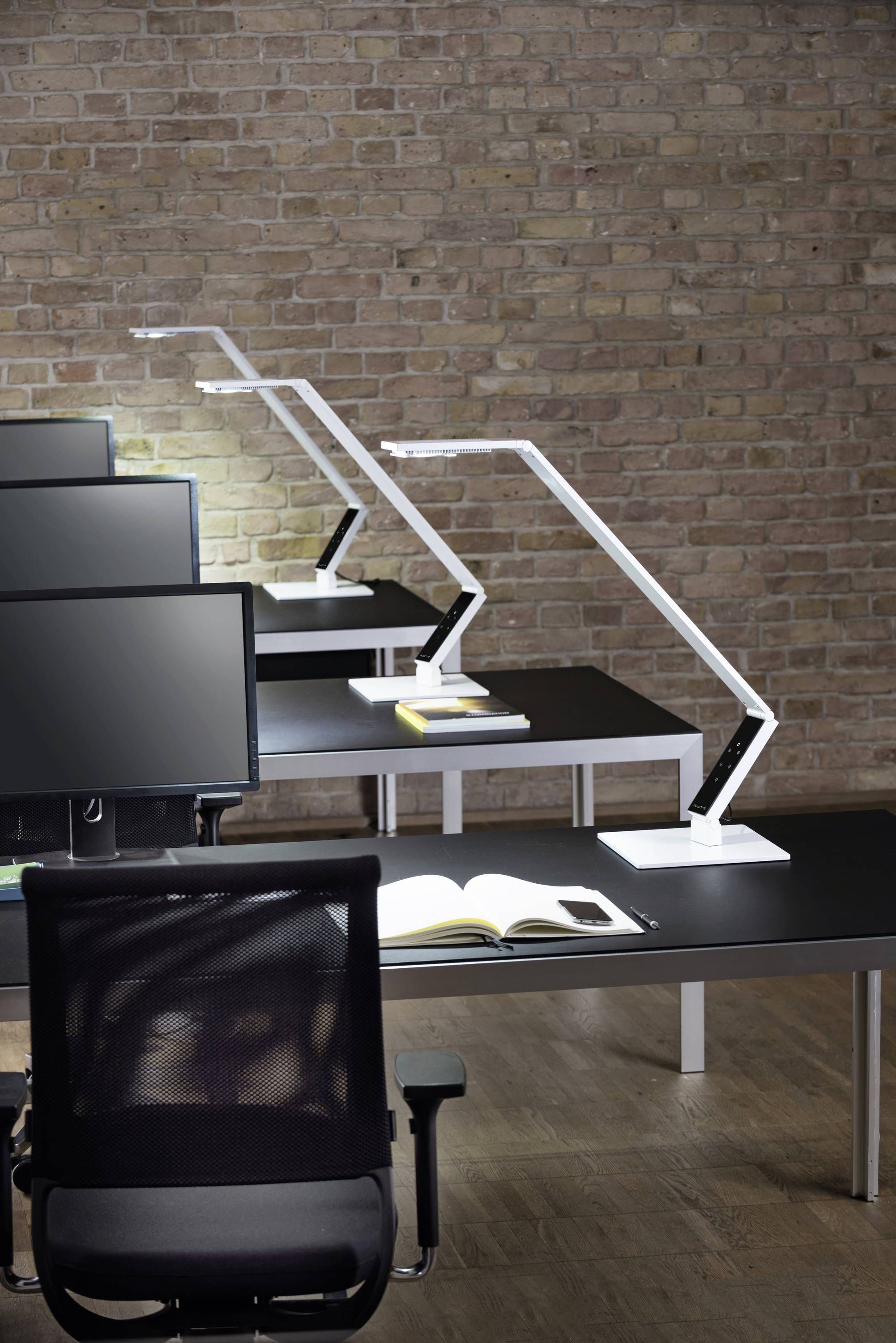 Modern office interior with black desks, white lamps and monitors in front of a brick wall. An open book lies on a table.