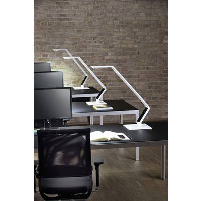 Modern office interior with black desks, white lamps and monitors in front of a brick wall. An open book lies on a table.