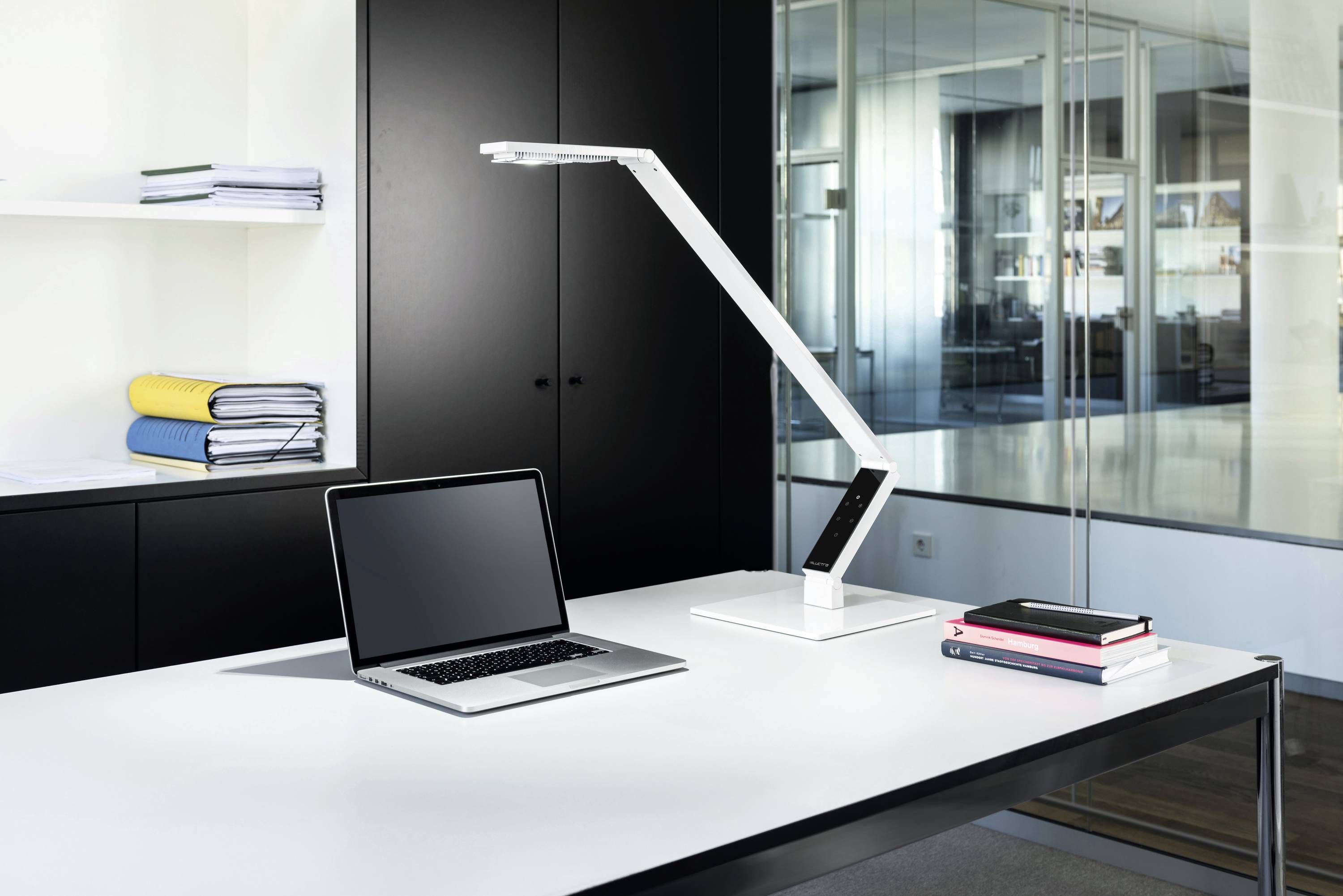 A modern desk in an office with a laptop, a white desk lamp, and stacked books on a white table. Filing cabinets and glass partitions are visible in the background.