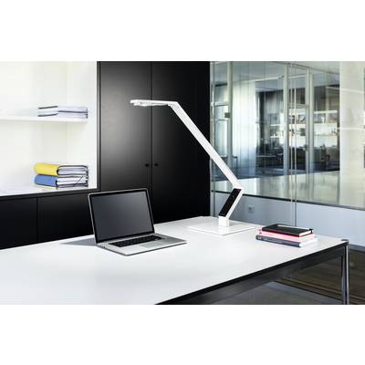 A modern desk in an office with a laptop, a white desk lamp, and stacked books on a white table. Filing cabinets and glass partitions are visible in the background.