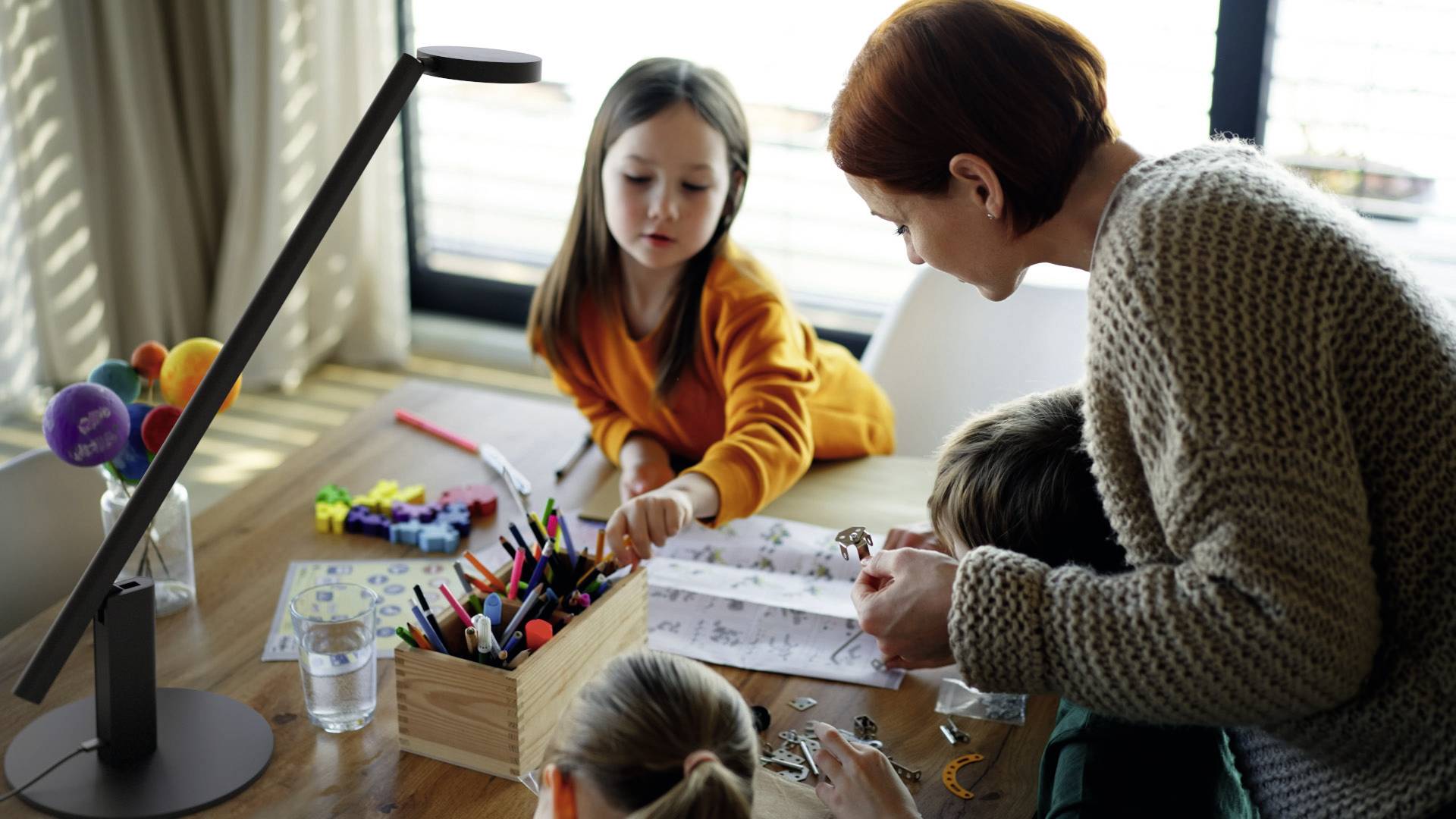 A woman and three children are sitting at a table crafting together. Craft materials and a container of pens are spread out on the table.