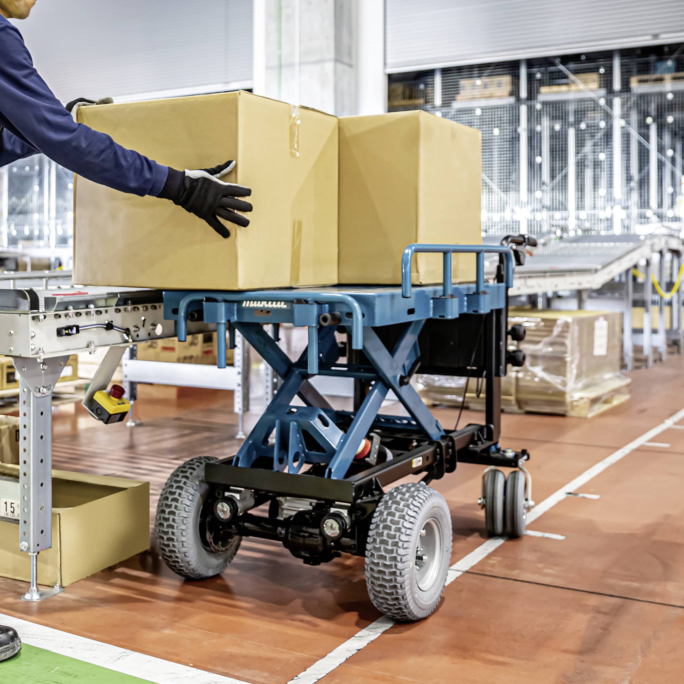 A worker is pushing a hydraulic pallet truck loaded with large boxes in a warehouse. Shelves and additional boxes can be seen in the background.