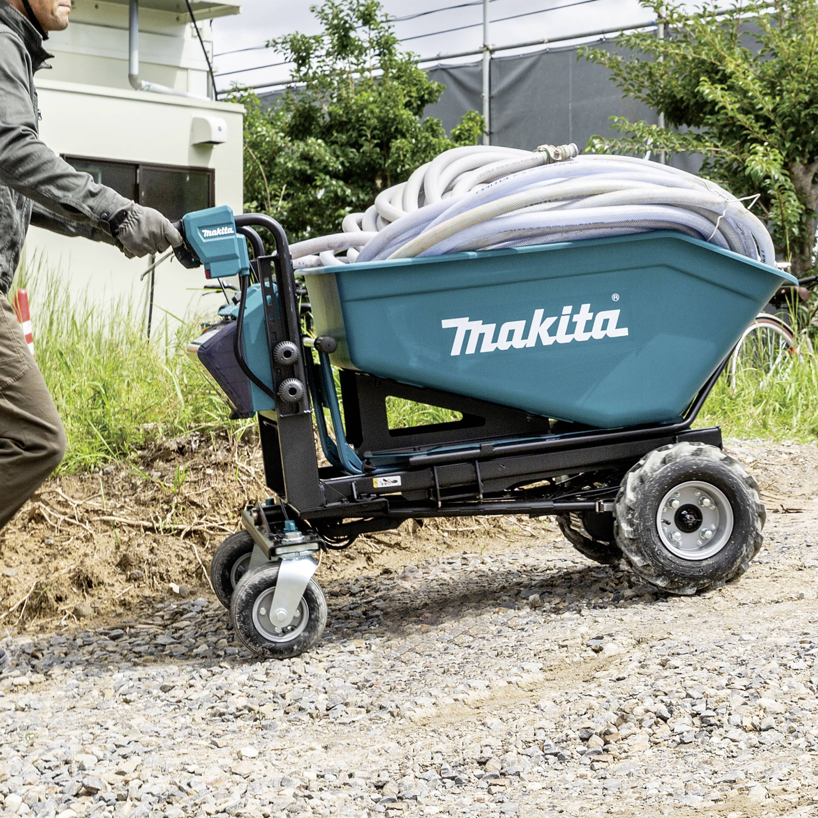 A person is pushing a motorised wheelbarrow by 'Makita' along a gravel path. Hoses are lying on the ground. Shrubs are visible in the background.