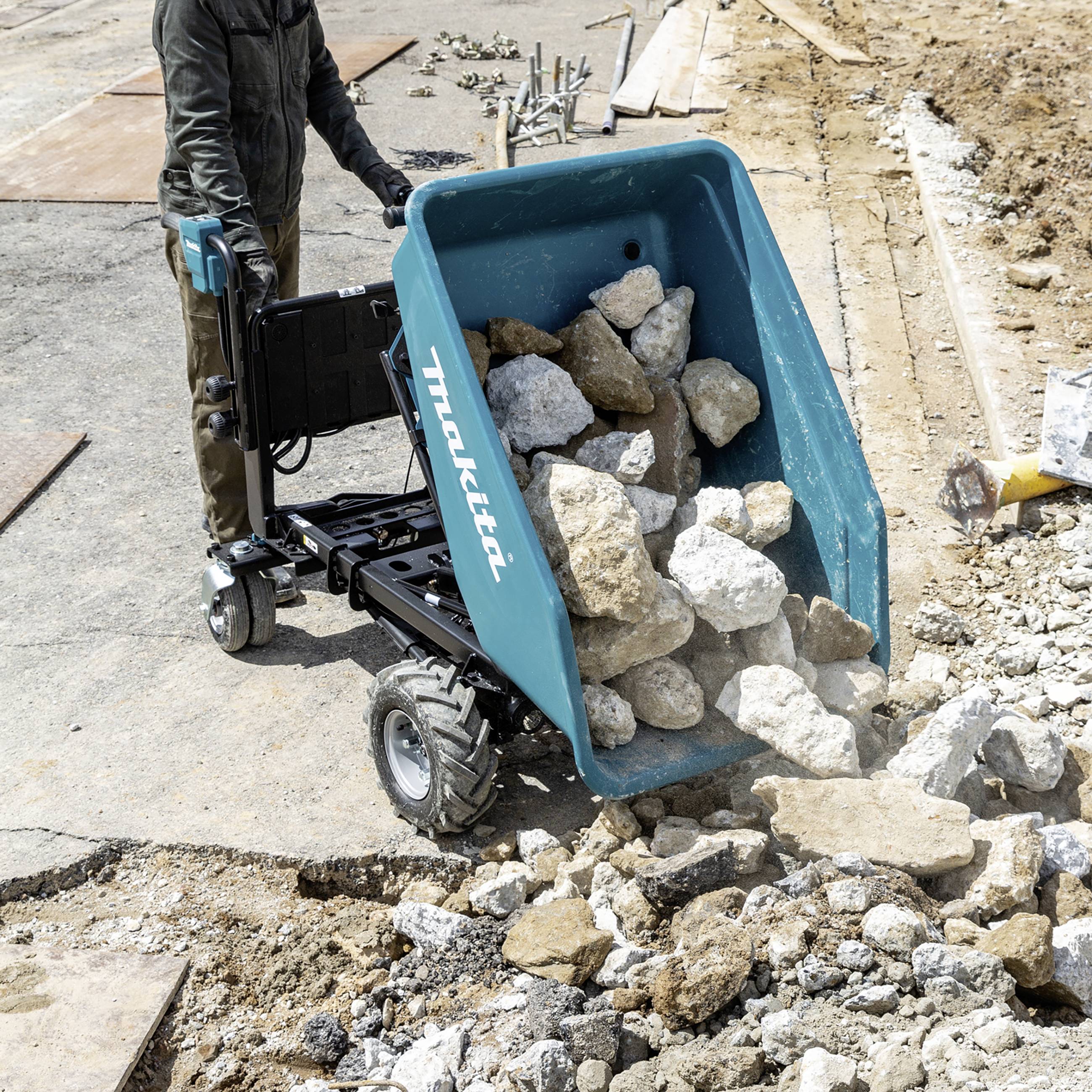 A person is operating an electric wheelbarrow laden with large stones on a construction site. The structure in the background indicates ongoing building work.