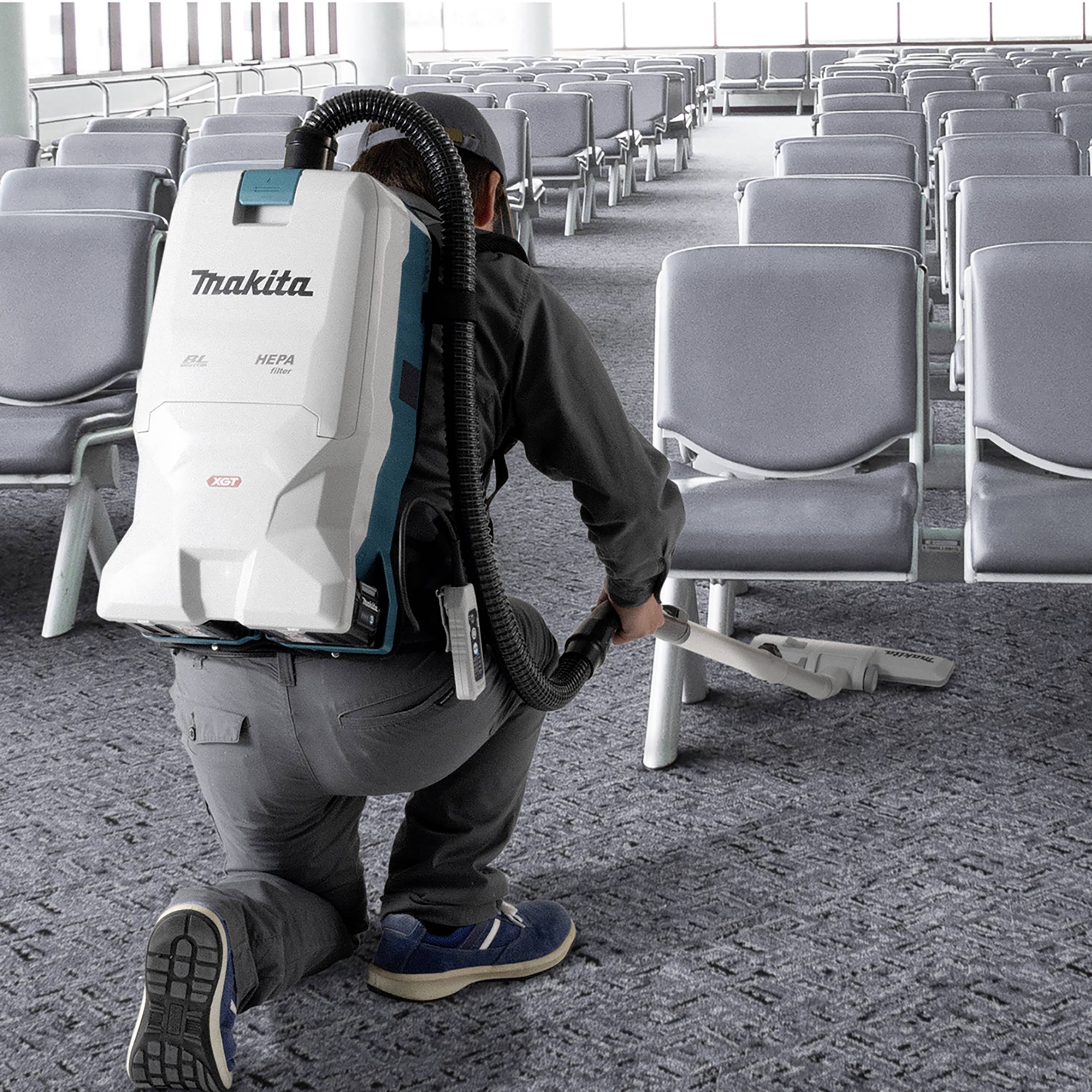 A person is cleaning a carpeted floor with a portable backpack vacuum cleaner in a large, empty conference room.