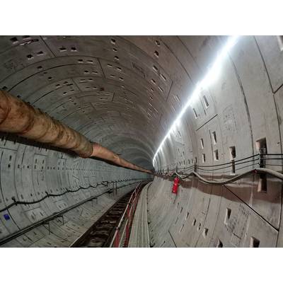 View into a concreted underground railway tunnel with lighting, pipes, and cables along the walls. A fire extinguisher is visible.