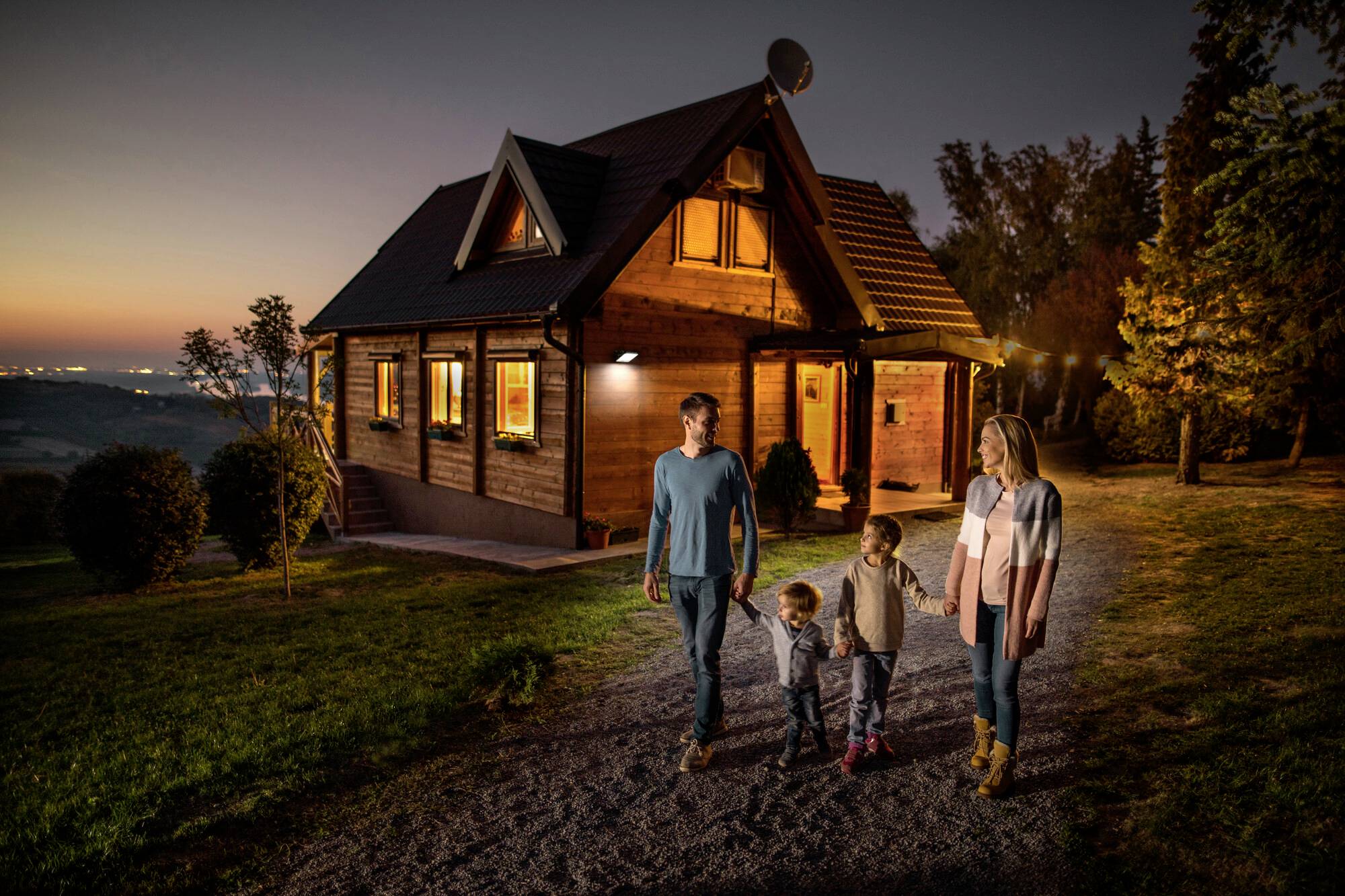 A family walks along a path at sunset in front of an illuminated wooden house.