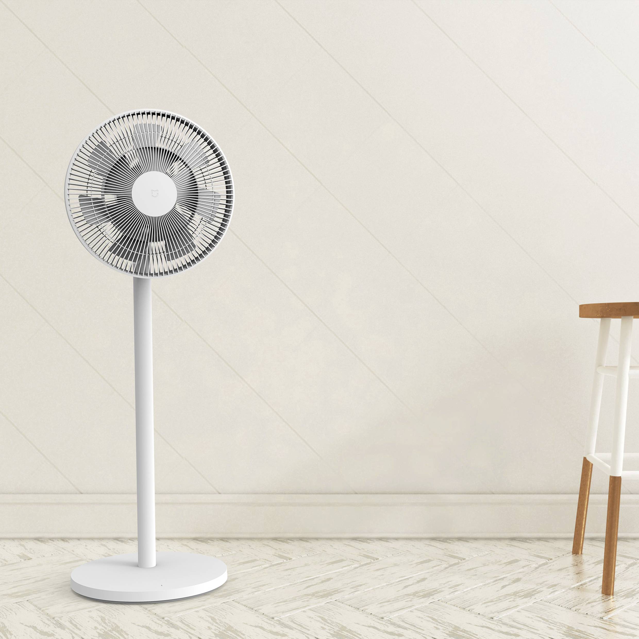 A white pedestal fan stands in a room with a wooden floor. Next to it is a small wooden table.