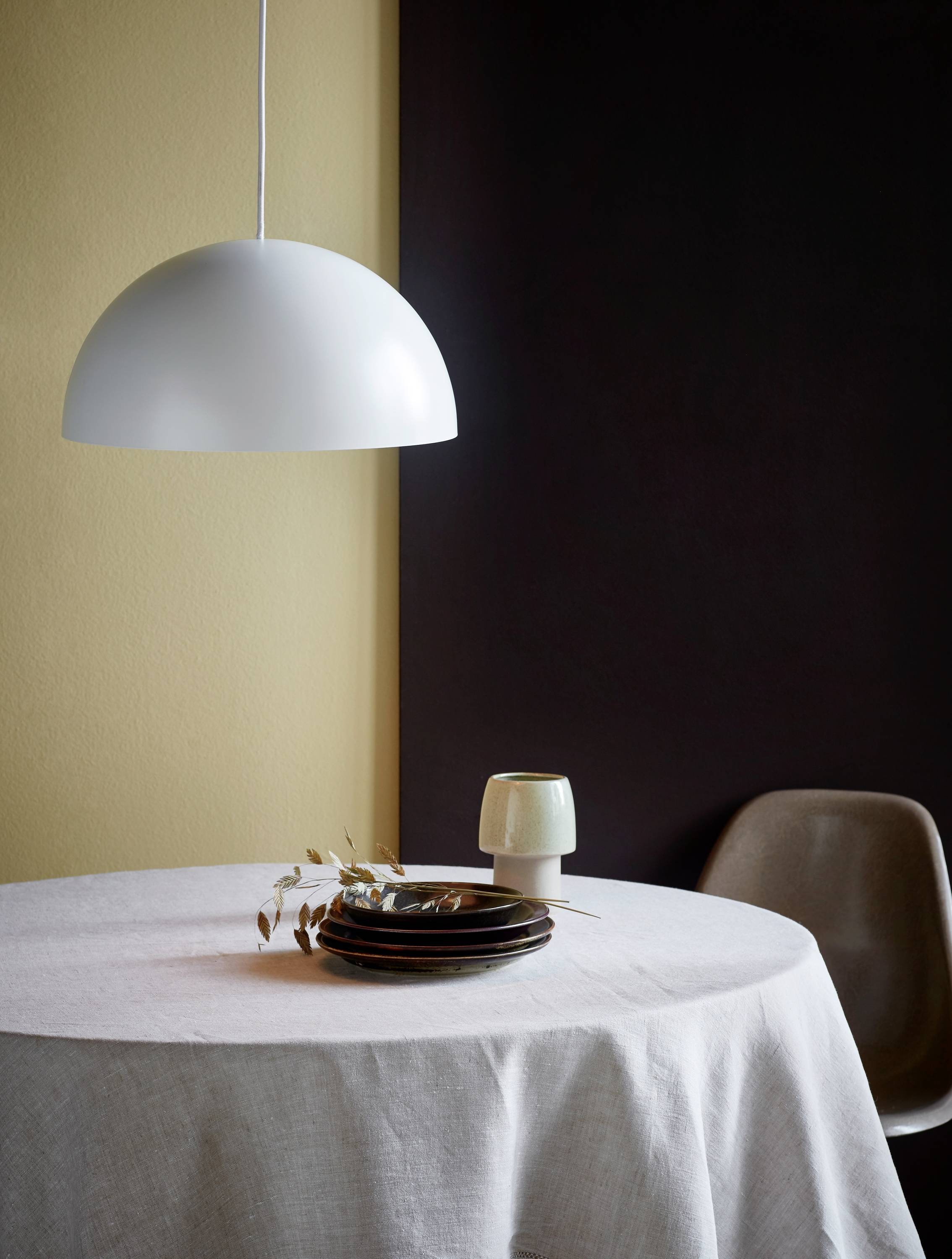A round dining table with a white tablecloth, on which are stacked plates, a white cup, and a white pendant light hanging from the ceiling.