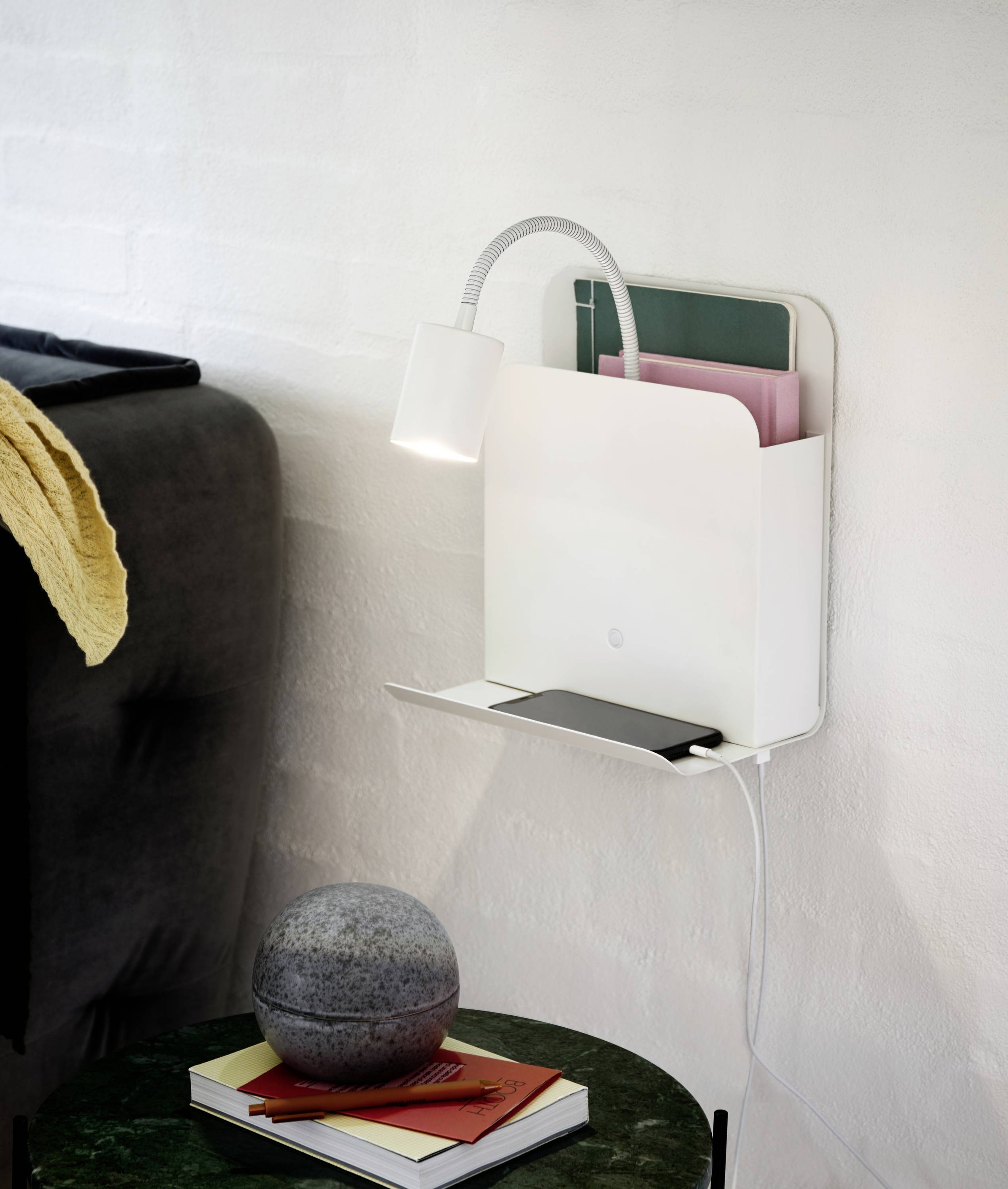 A white wall light with a flexible reading lamp and a built-in shelf that holds books and a mobile phone, positioned next to a sofa.