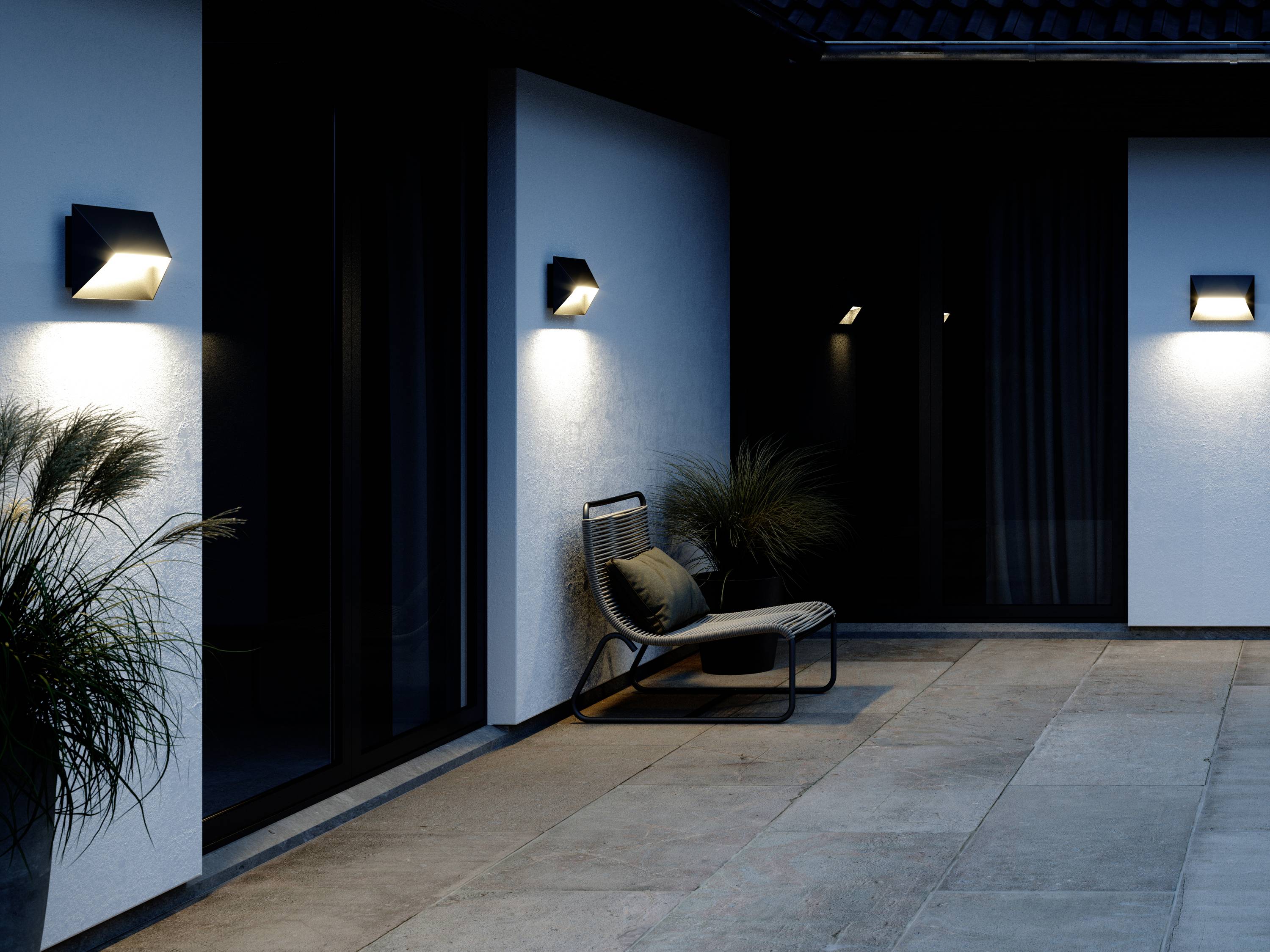 A modern, illuminated outdoor space with stone flooring, plants, and a lounger against a house wall at dusk.