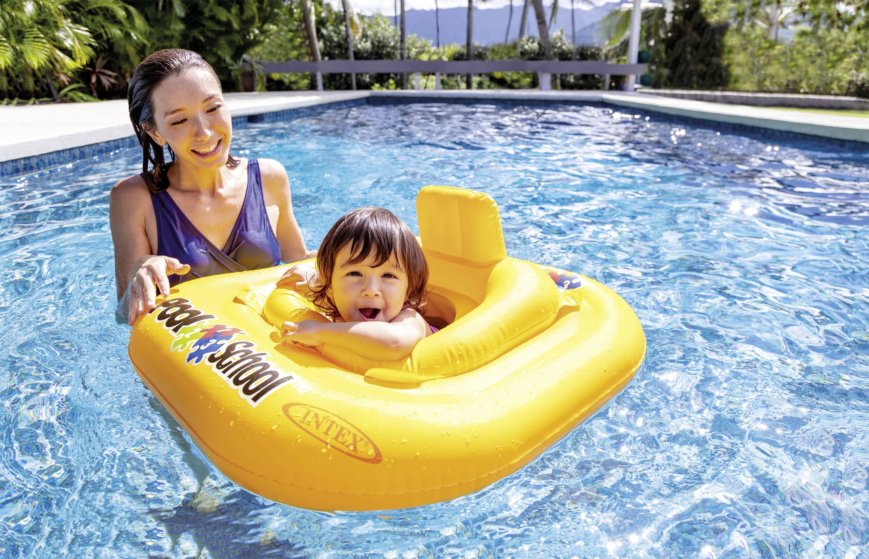 A woman stands in a swimming pool, accompanying a small child sitting in a yellow rubber ring. Trees can be seen in the background.