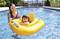 A woman stands in a swimming pool, accompanying a small child sitting in a yellow rubber ring. Trees can be seen in the background.