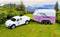 A white pickup truck is pulling a pink horse trailer across a meadow, surrounded by trees and hills in the background.