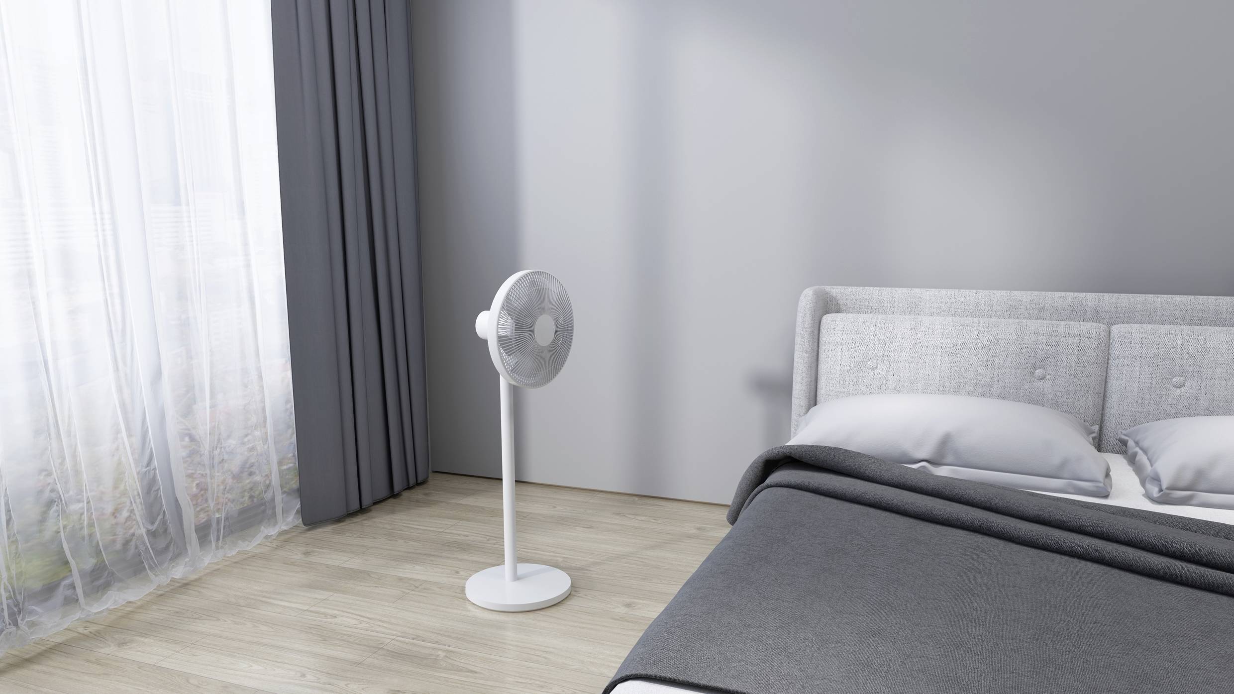 A grey bedroom with a standing fan next to a bed with grey bedding, on a light wooden floor.