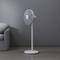 A white pedestal fan stands on a wooden floor next to a grey section of sofa. The fan casts a shadow on the wall.