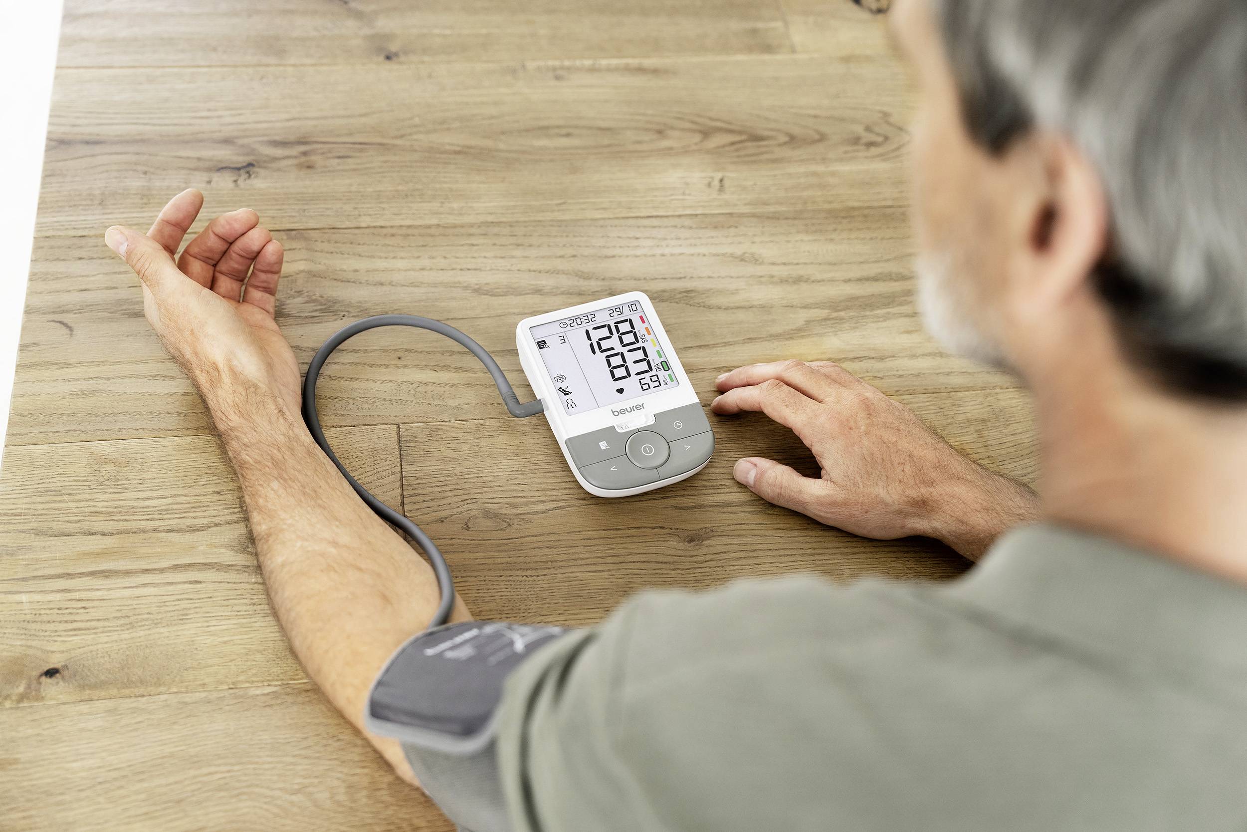 A man is measuring his blood pressure with a digital blood pressure monitor on a wooden table. The display is showing readings.