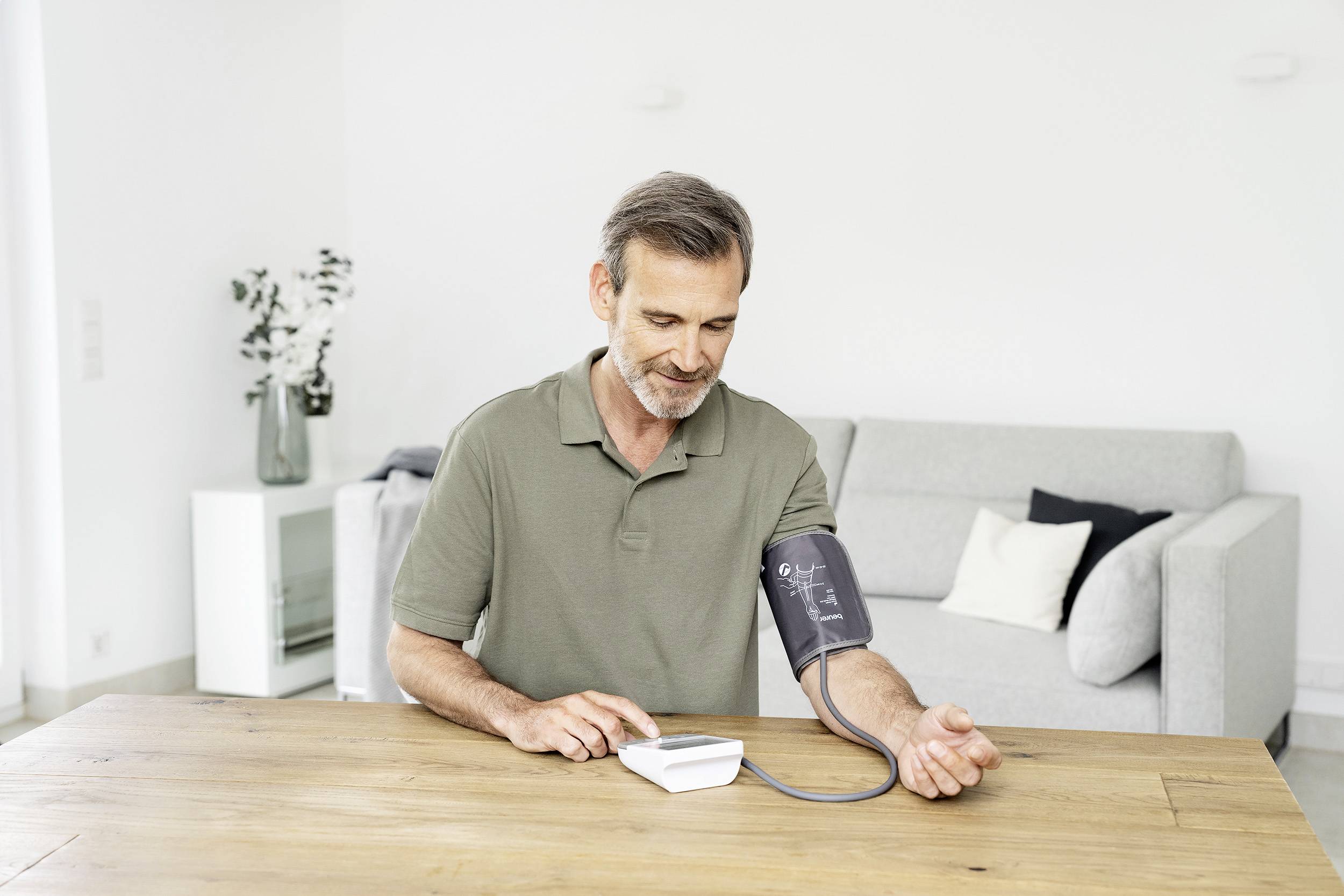 A man is measuring his blood pressure with a digital device at a table in a bright living room. A sofa is visible in the background.
