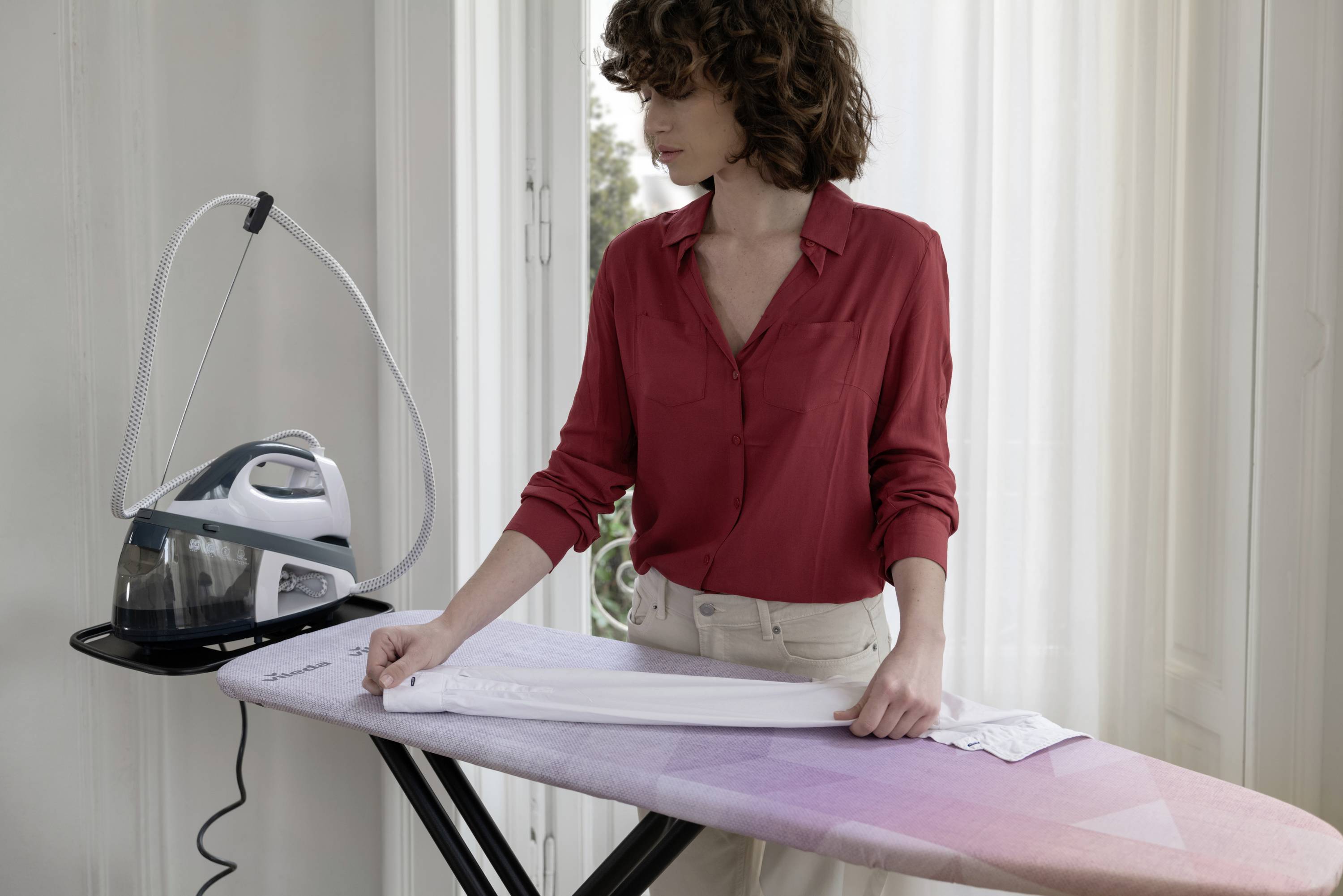 A woman is ironing a garment on an ironing board in a bright room. A steam iron is standing next to her.