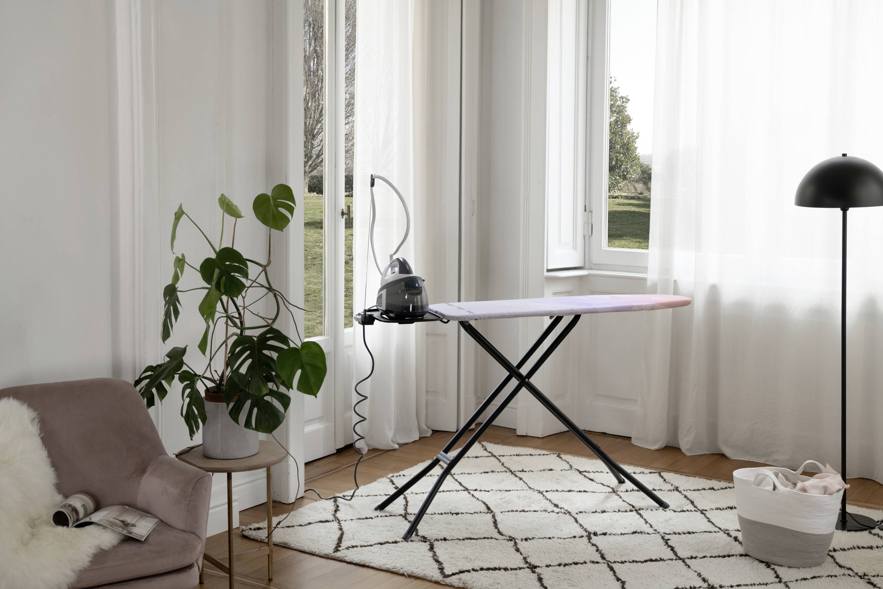 A bright room with an ironing board in the centre, beside which stands a plant stand. Large windows allow plenty of light to flood in.
