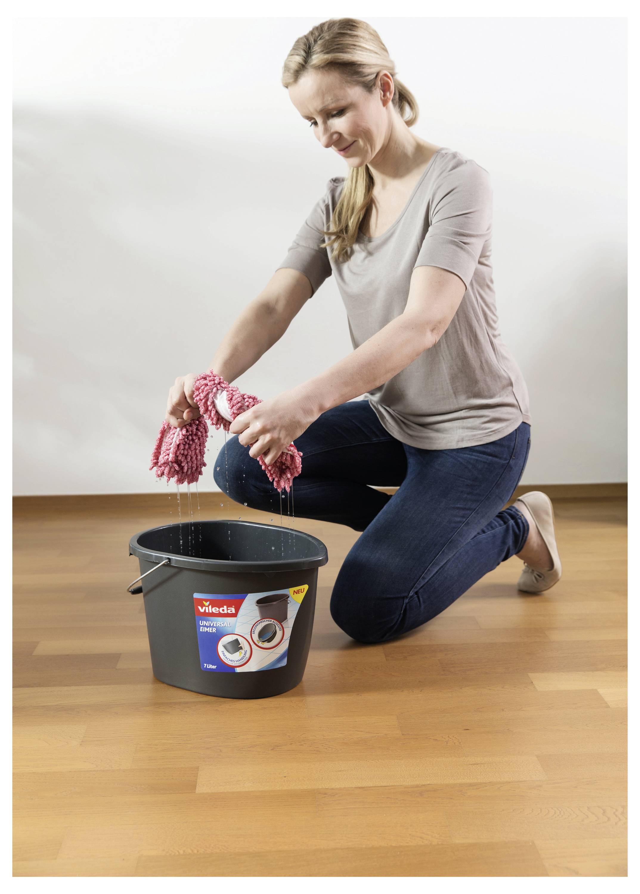 Woman kneeling on a wooden floor wringing a wet mop over a bucket, cleaning the floor.