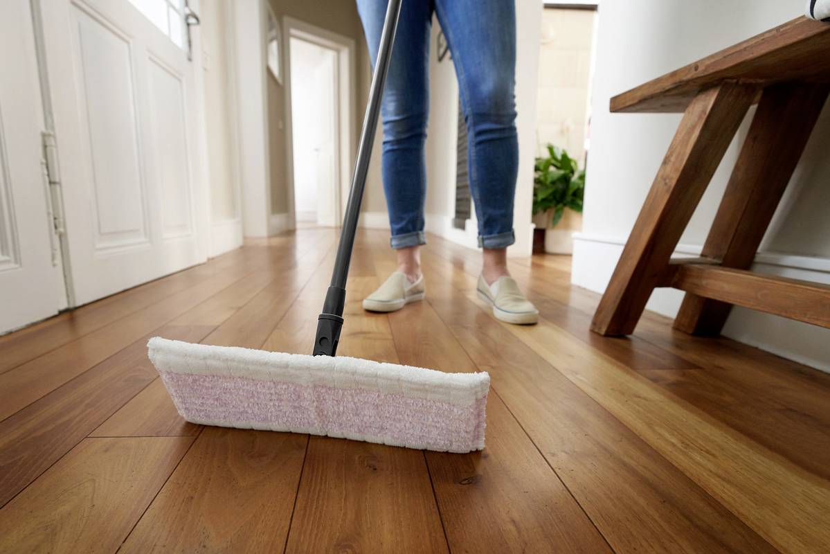 A person is mopping the wooden floor of a hallway with a flat mop. An open door and a plant are visible in the background.