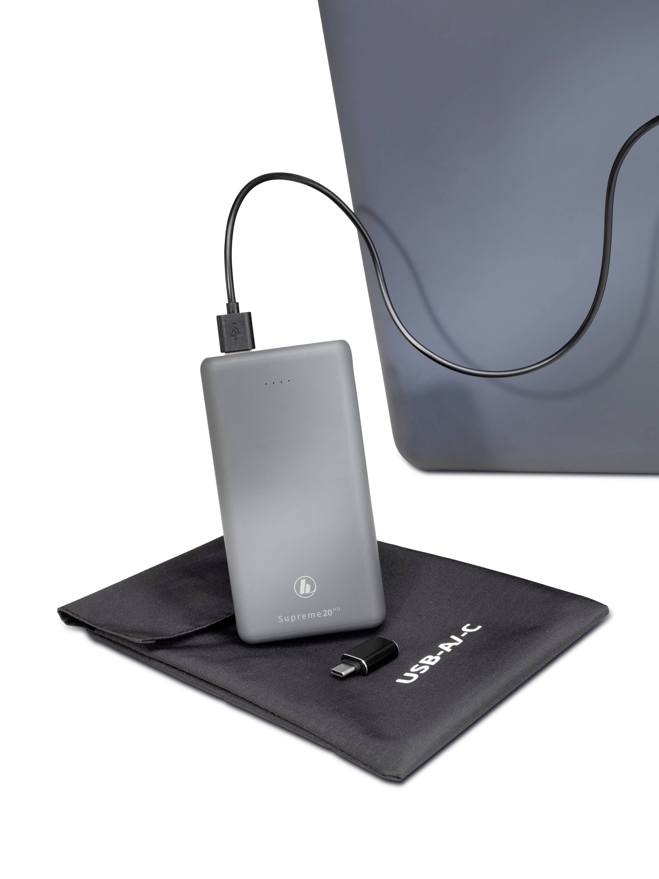 A silver power bank is connected to a laptop via a cable. Underneath the power bank is a black bag with the inscription 'USB-C'.