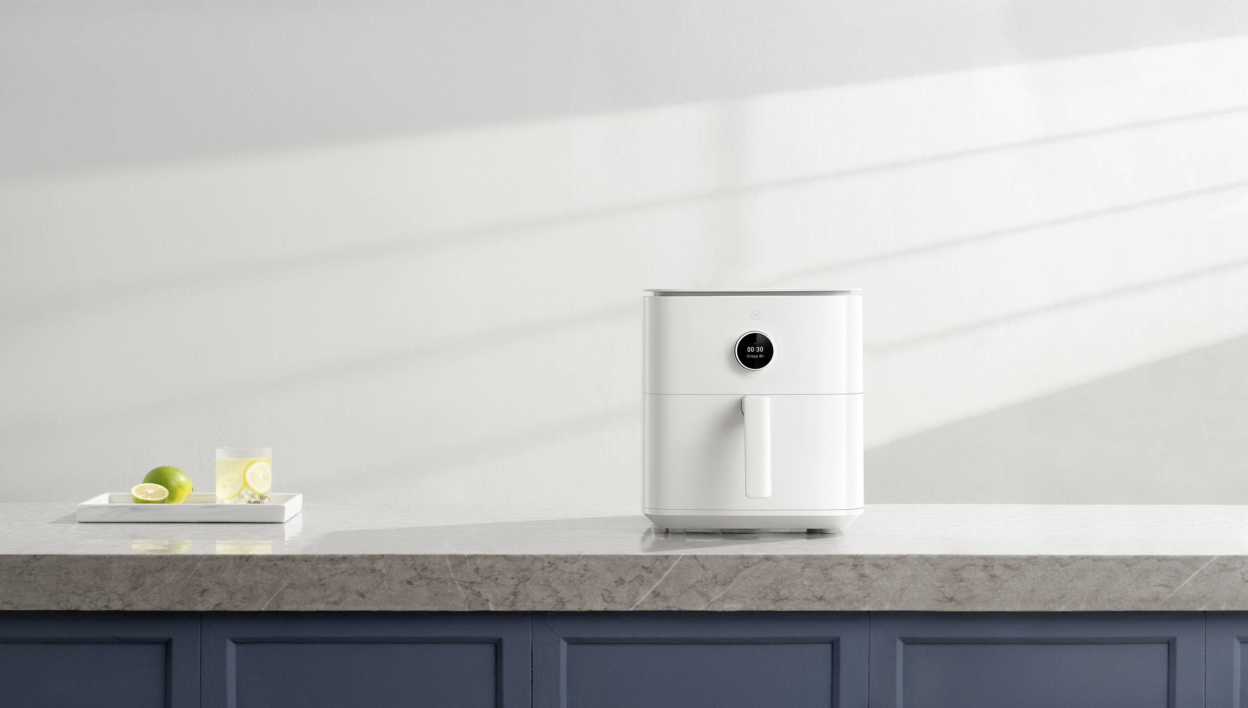 White hot air fryer on a kitchen countertop, beside a tray with a glass of water and lime slices. Modern kitchen setting.