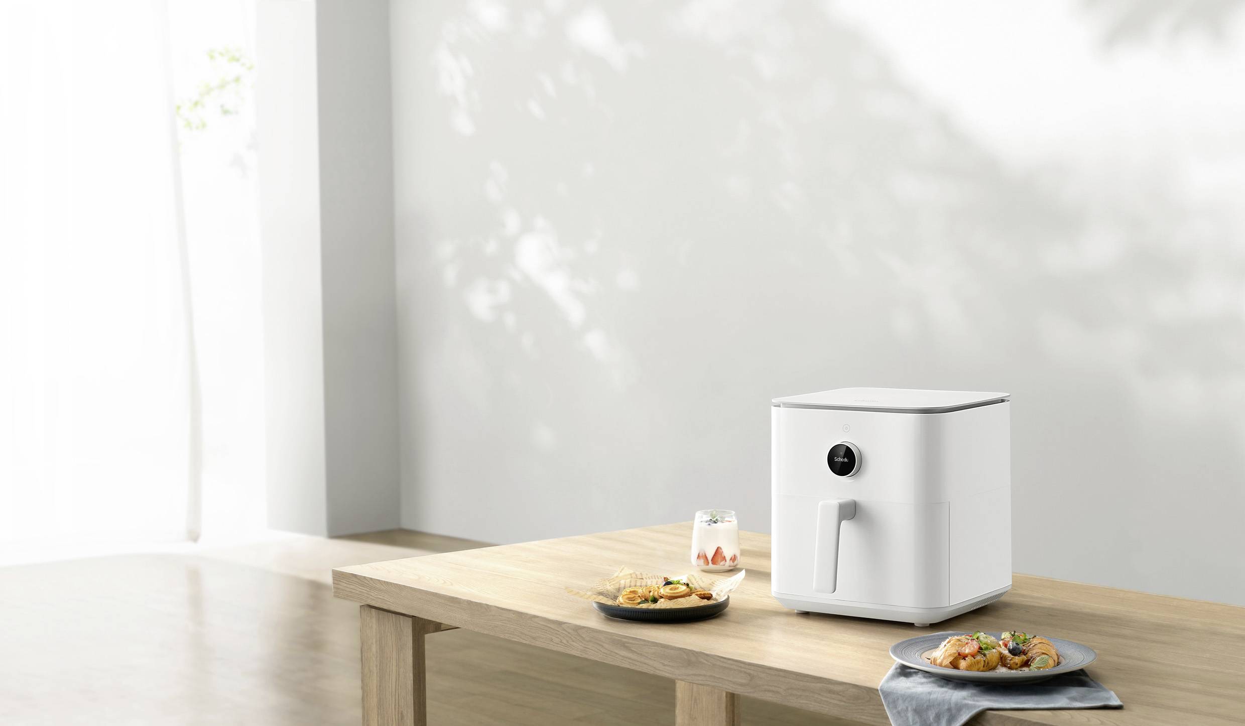 A white air fryer sits on a wooden table next to two plates of prepared food and a glass. Light streams through a window.