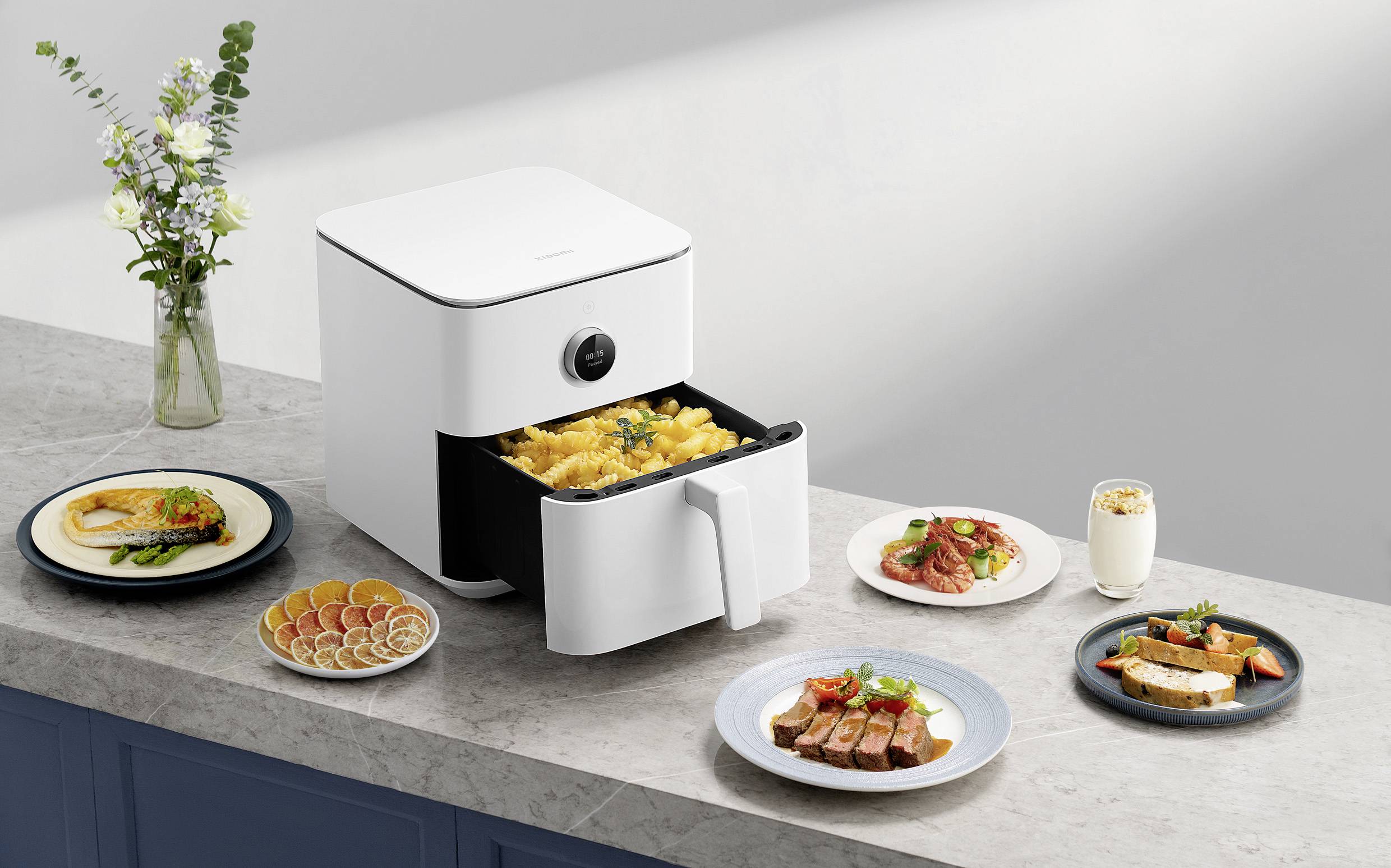 A white air fryer on a kitchen worktop is preparing pasta. Beside it are plates with various dishes and a drink.