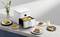A white air fryer on a kitchen worktop is preparing pasta. Beside it are plates with various dishes and a drink.